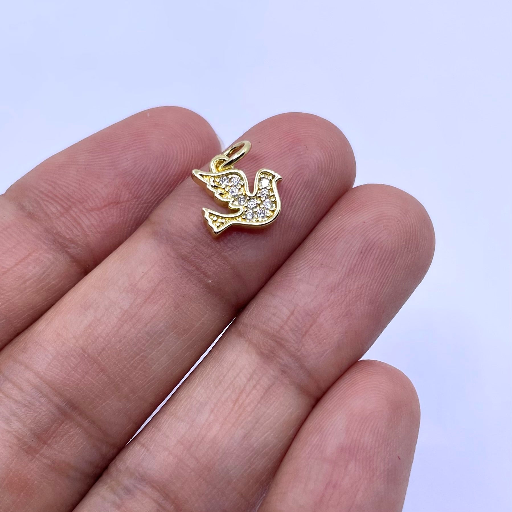 A small gold bird charm paved with clear stones, held between a person's fingers to show its size against a light blue background.
