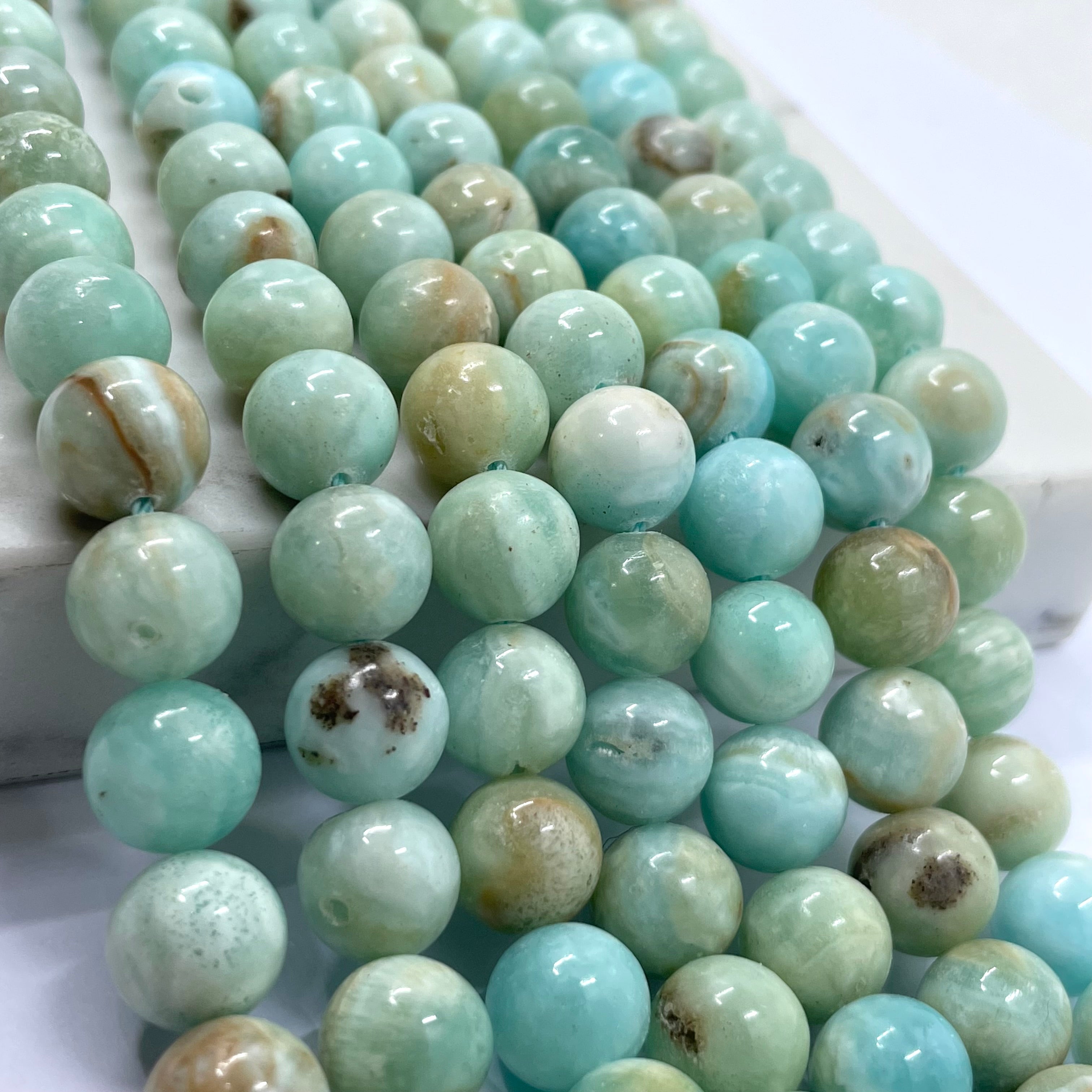 Close-up of strands of round light blue-green hemimorphite gemstone beads with natural marbling and a few brown inclusions, arranged on a white surface.