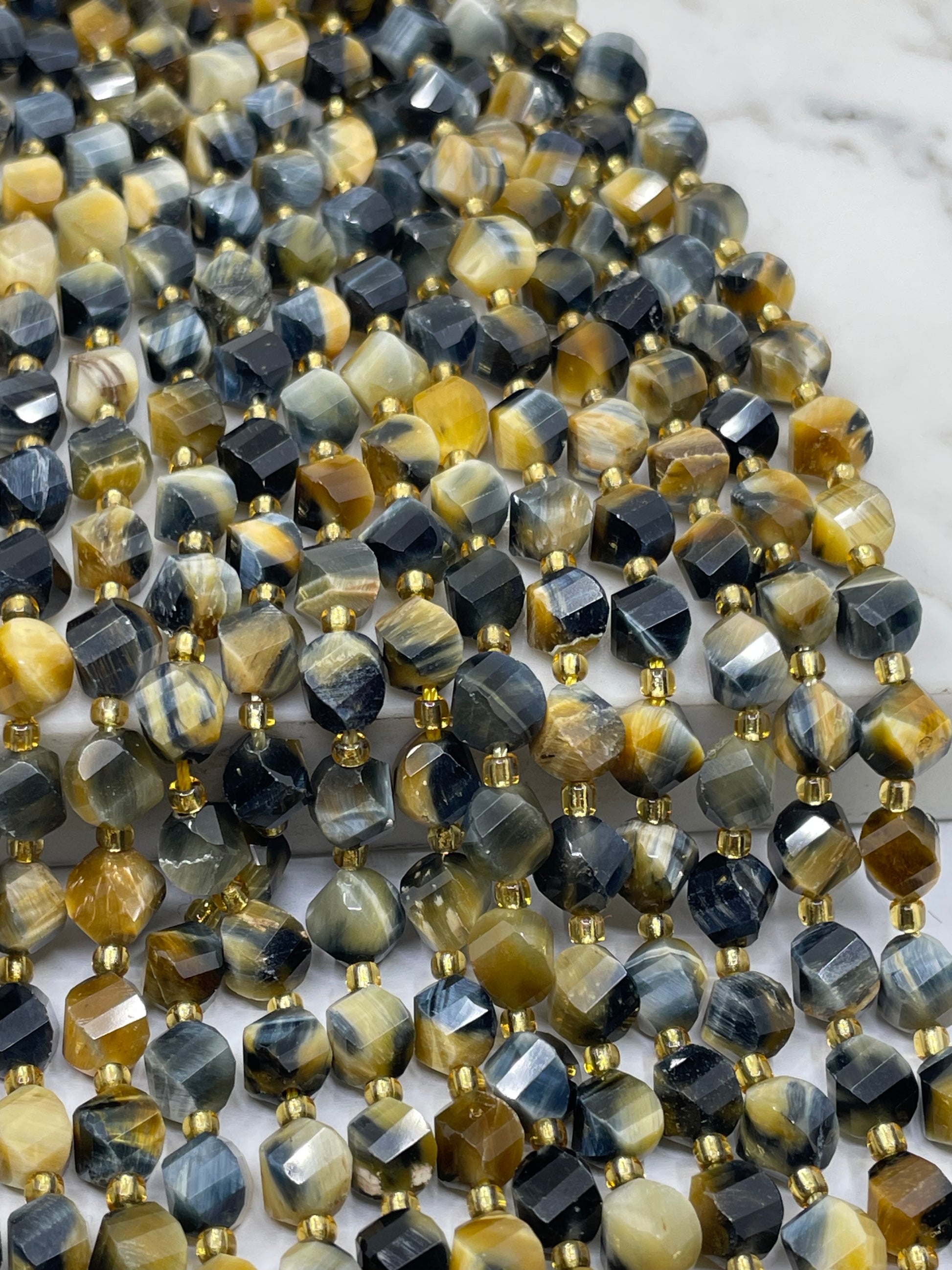 Close-up of faceted tiger eye beads strung in multiple strands, featuring tiger's eye patterns with black, gray, brown, and golden-yellow tones separated by small gold spacer beads, set against a light marble background.
