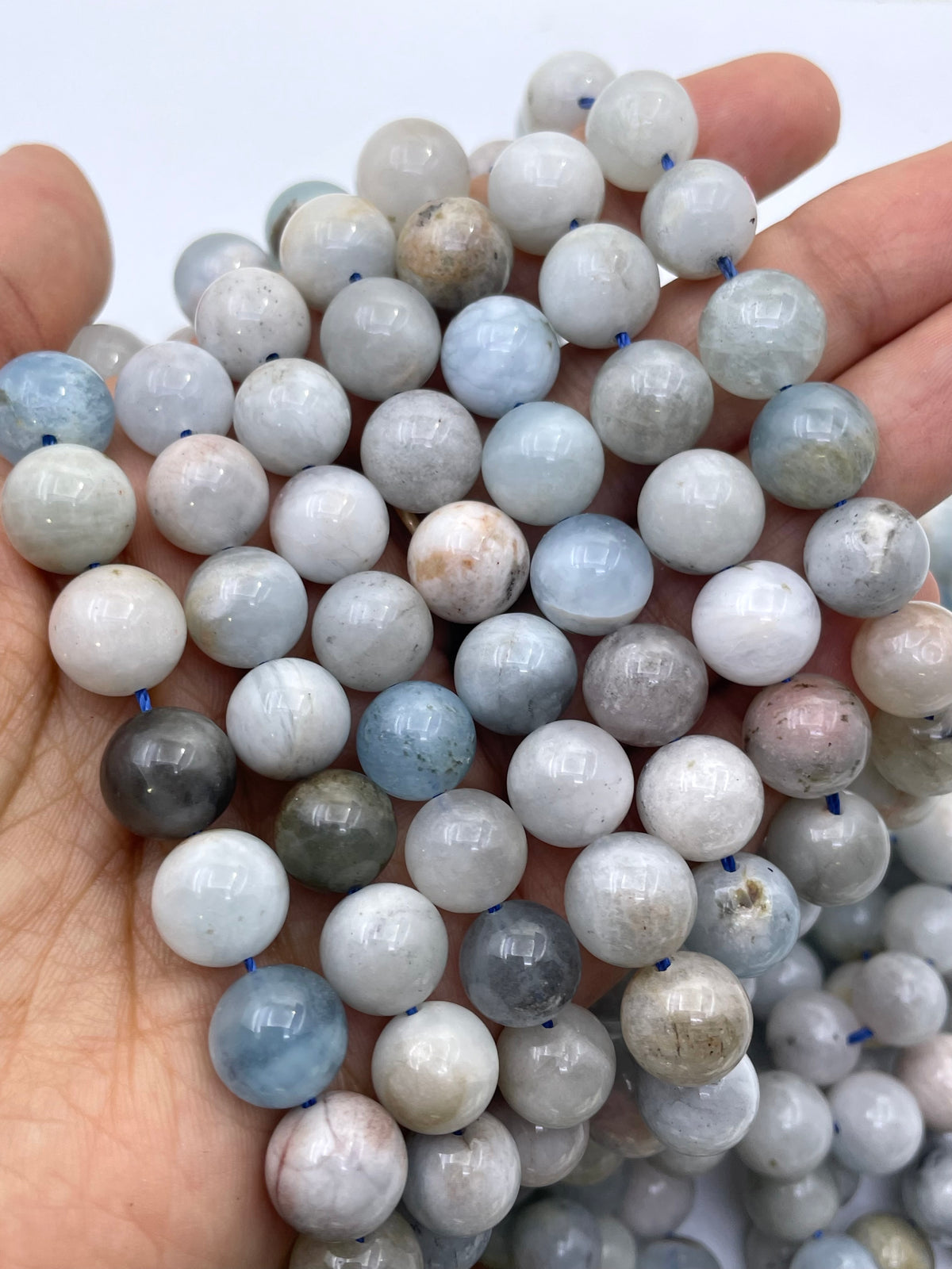 A hand holding a pile of round, polished aquamarine gemstone beads in light blue, gray, white, and soft pink tones, strung on thin blue cords.