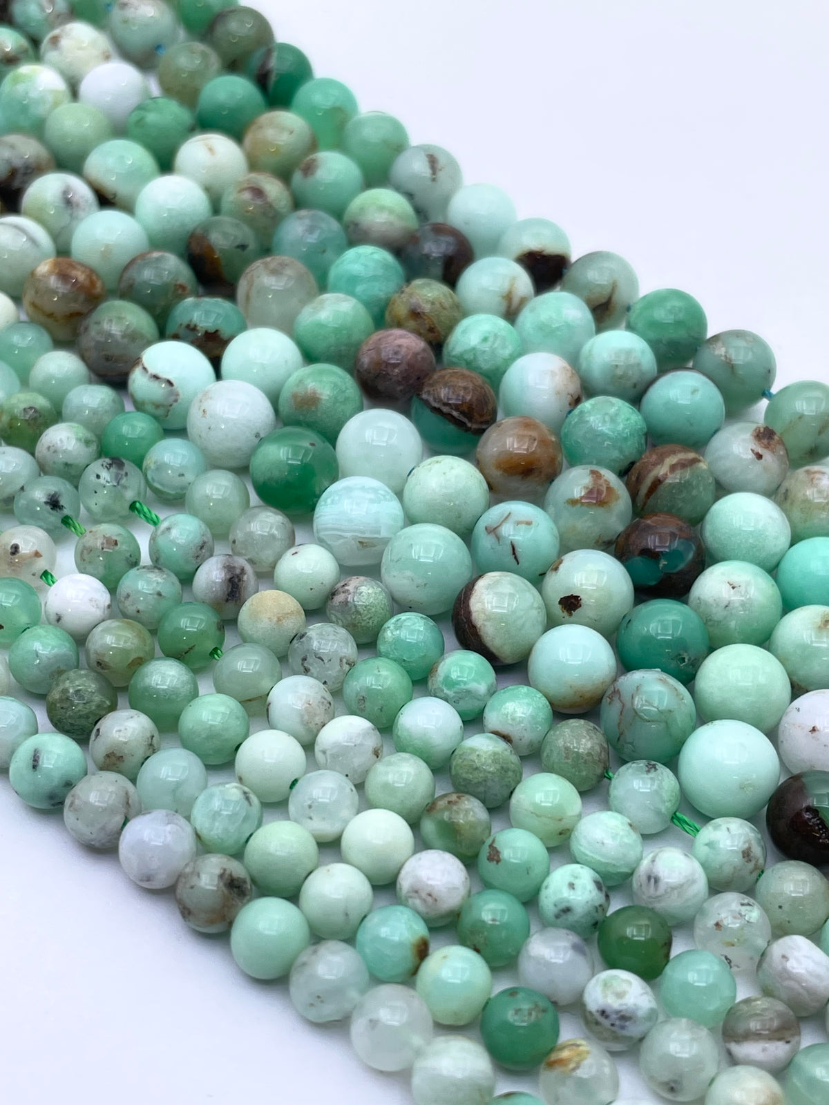 Faceted round gemstone beads in various shades of light green, mint, and white with some natural brown matrix, arranged in a diagonal line on a white background.