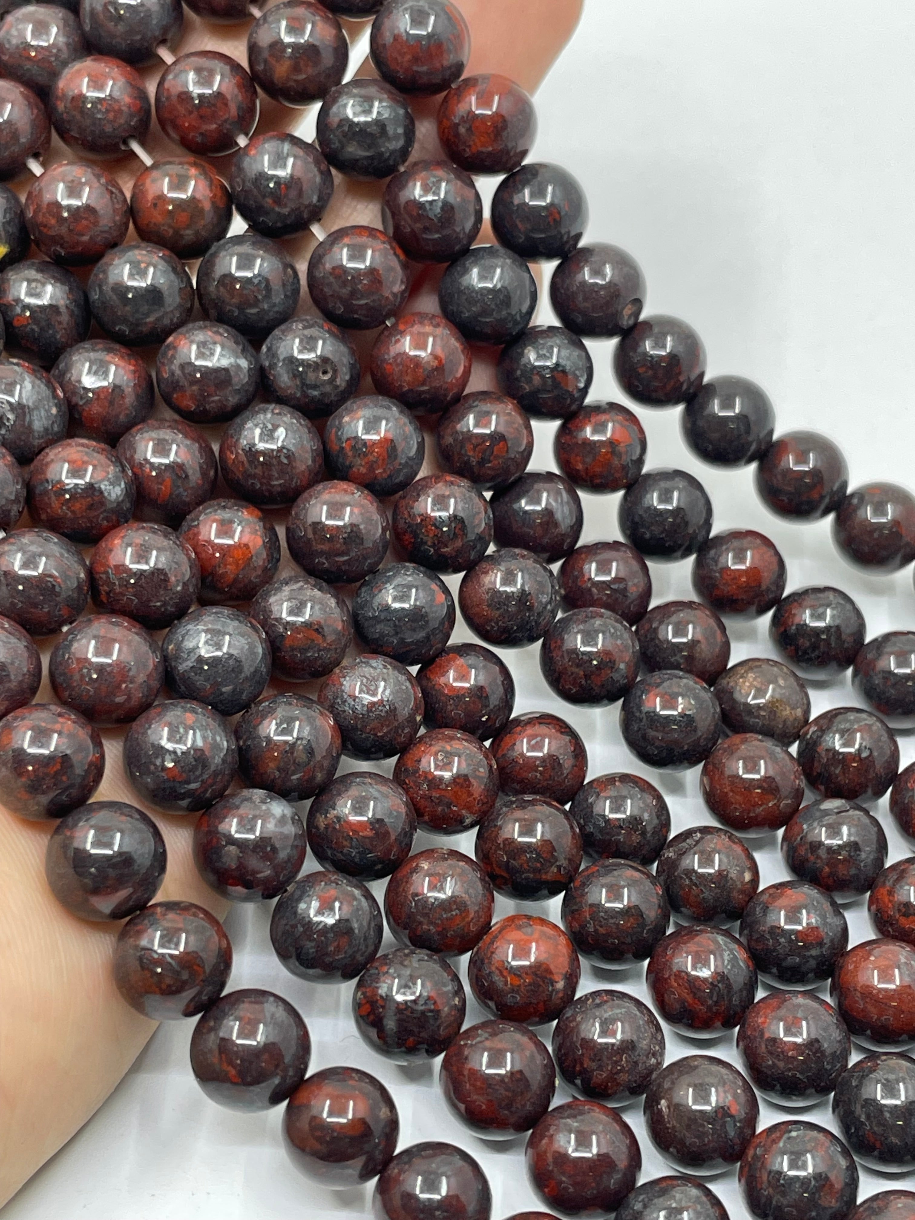 A close-up of glossy round brecciated jasper beads in a dark reddish-brown and black marbled pattern, held in a hand against a light background.