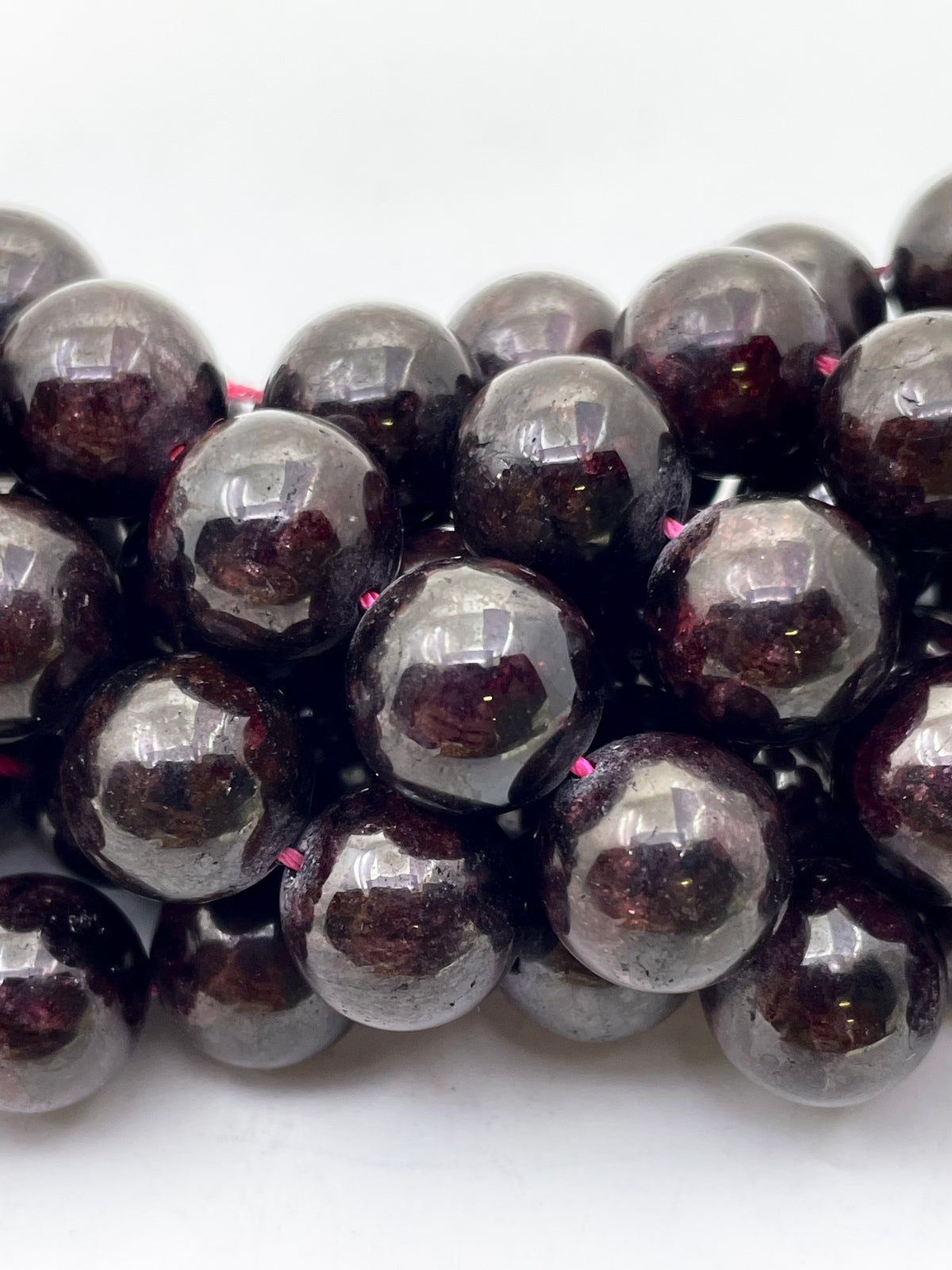 A close-up of glossy garnet beads with a deep purple-red internal inlay, strung on a thin pink cord against a plain white background.