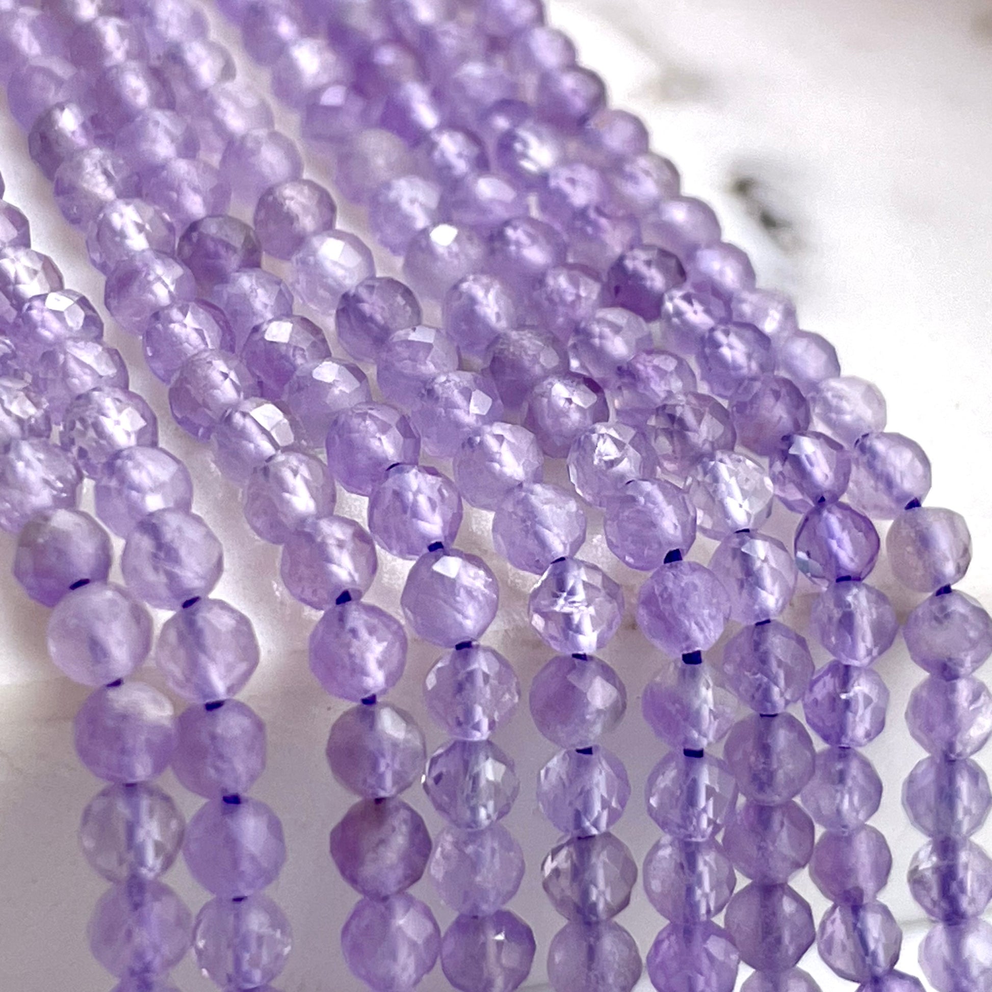 Faceted lavender purple gemstone beads strung on multiple strands, displayed on a light marble-like surface.
