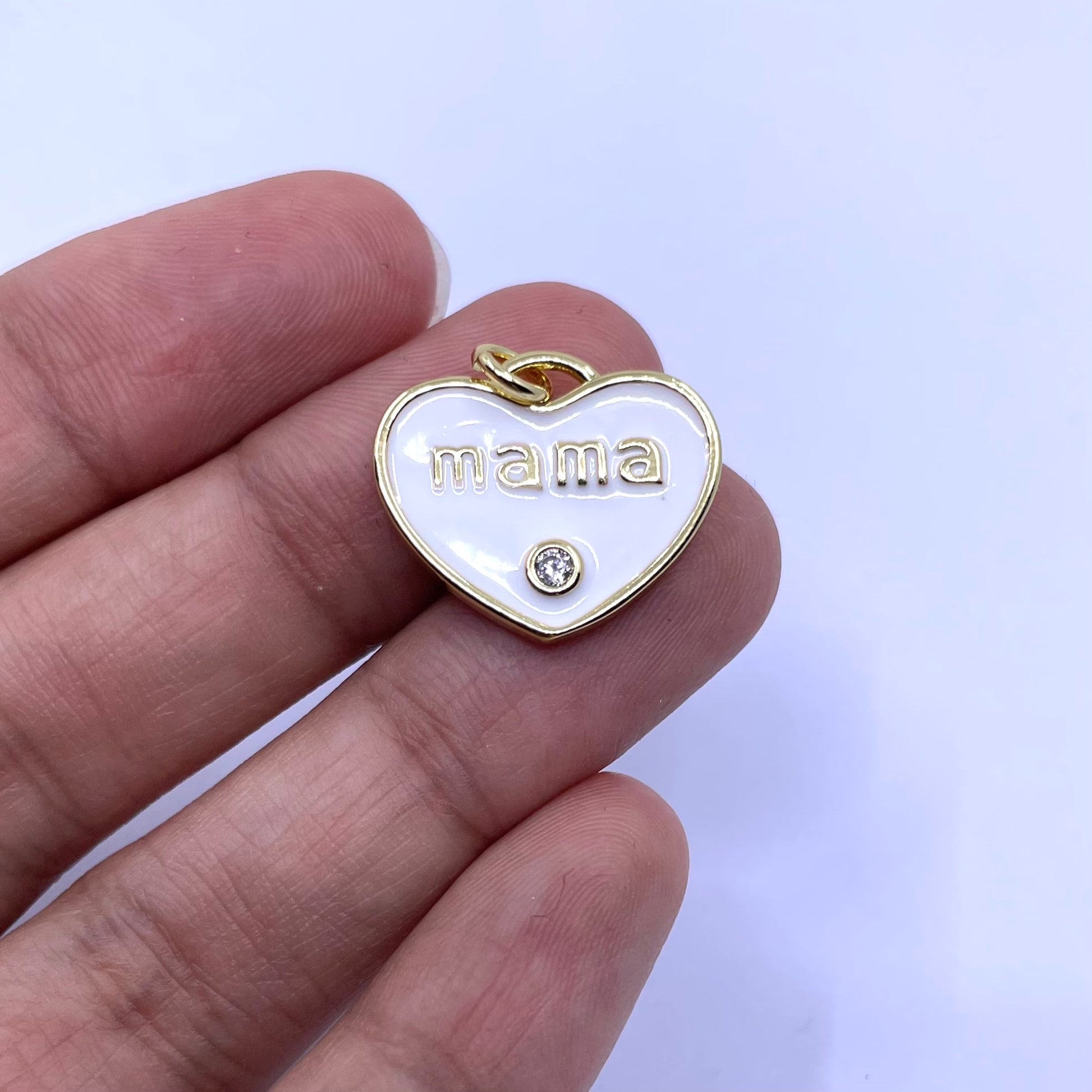 A gold heart-shaped pendant with a white enamel background, featuring the word 'mama' in gold lowercase letters and a small clear rhinestone below it, shown resting on a person's fingers against a light background.
