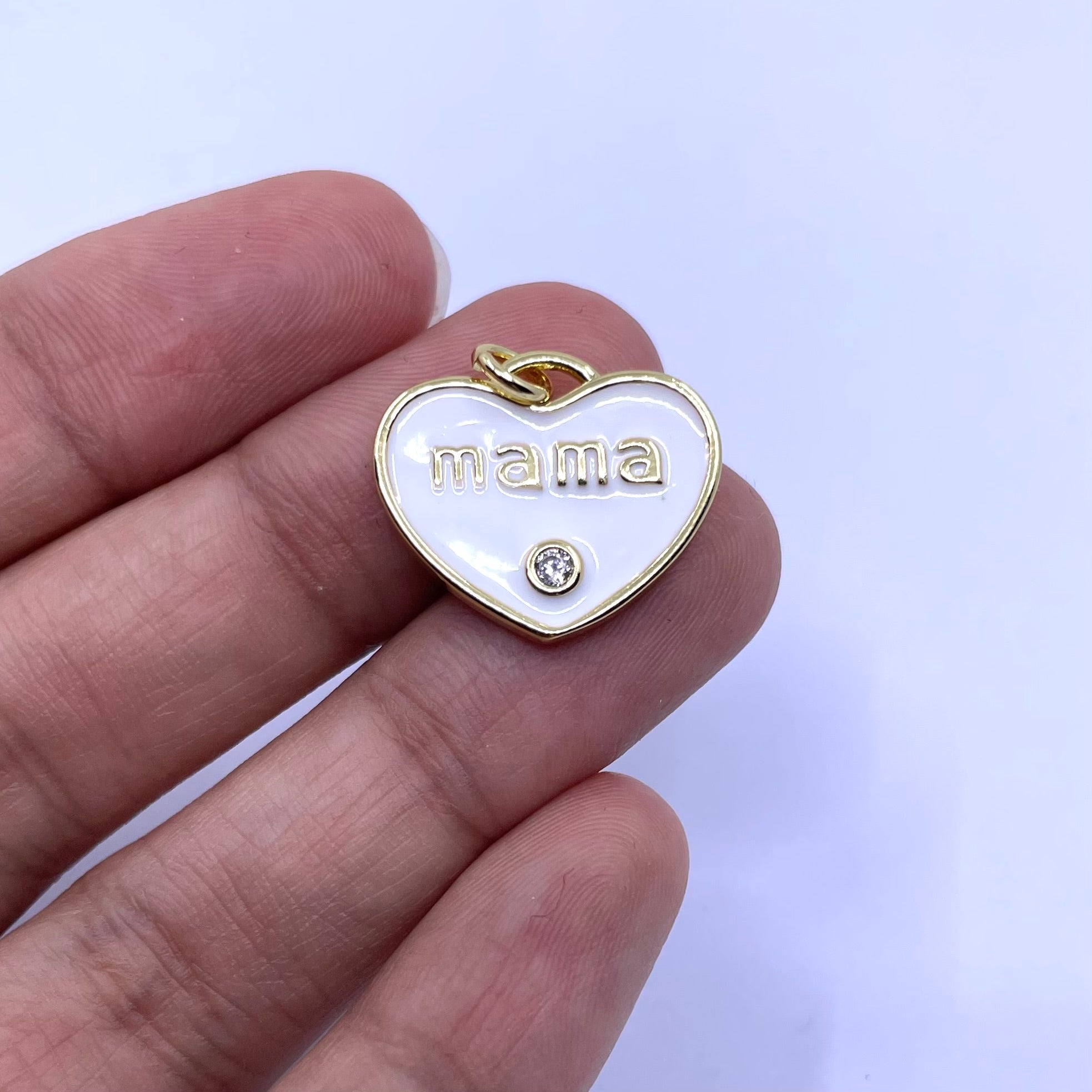 A gold heart-shaped pendant with a white enamel background, featuring the word 'mama' in gold lowercase letters and a small clear rhinestone below it, shown resting on a person's fingers against a light background.