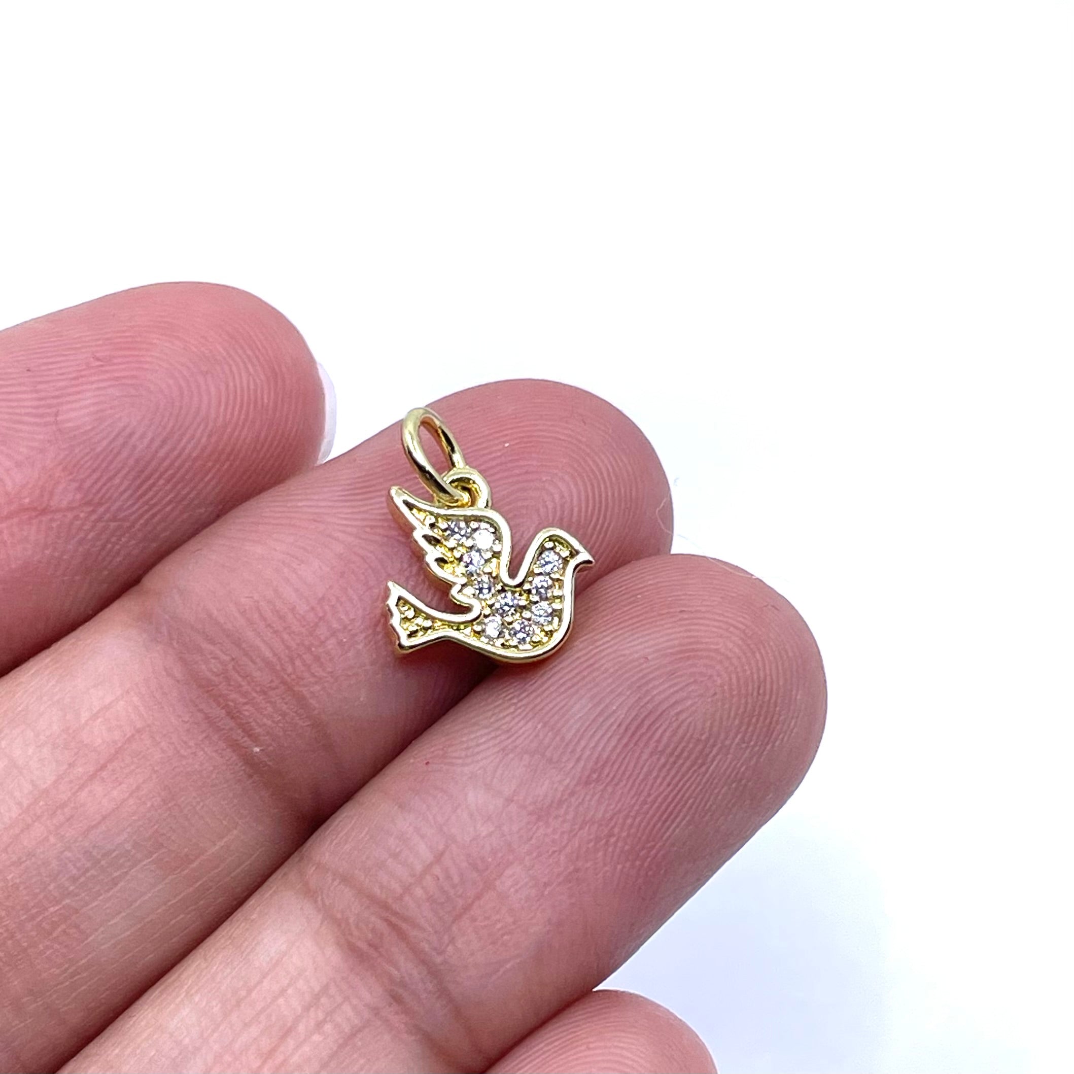 A small gold dove-shaped pendant with clear stones, shown resting on a person's fingers against a white background.