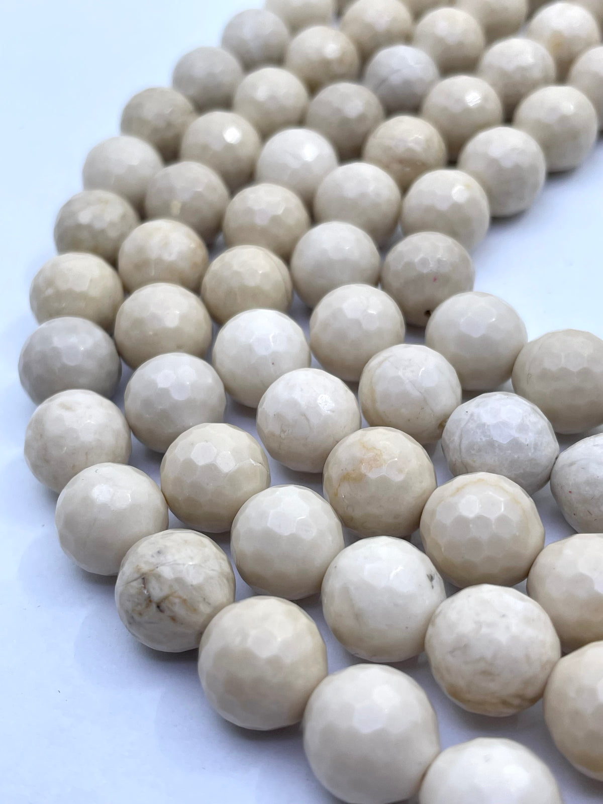 Faceted round riverstone/ fossil stone beads in a warm cream or ivory tone, arranged in curved rows on a plain light background.