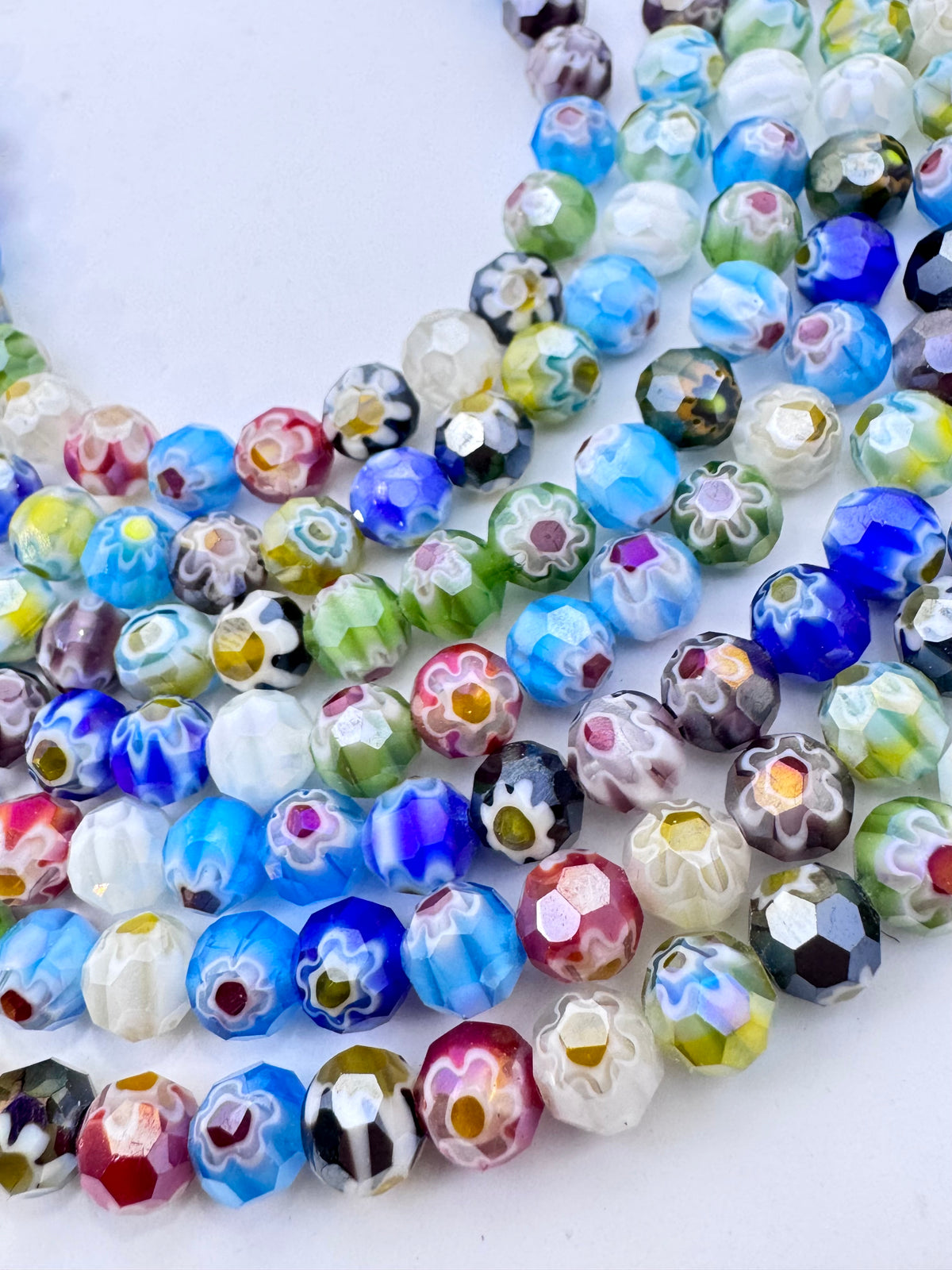 A close-up of many faceted glass beads arranged in curved strands on a white background. The beads are multicolored with swirls and floral patterns in blue, white, pink, green, yellow, black, and purple.
