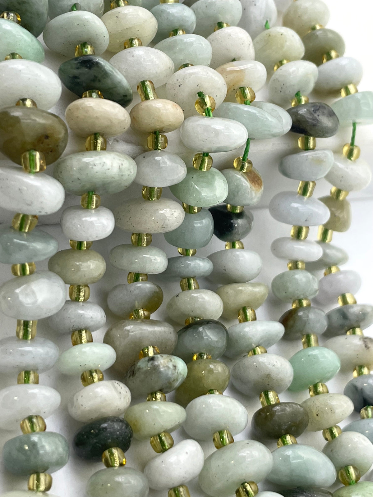 Multiple strands of polished burma jade stone beads in soft green, gray, white, and beige tones, separated by small metallic gold spacer beads, arranged closely together on a white background.