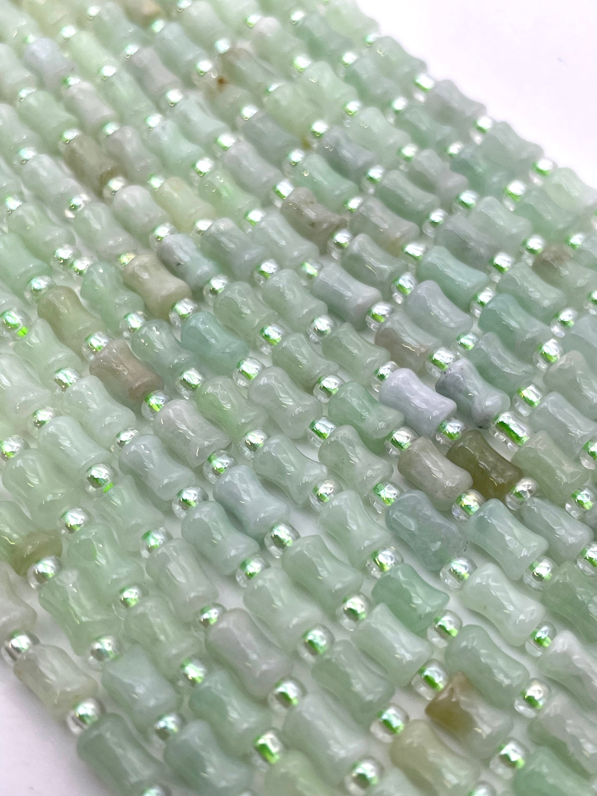 Close-up of multiple strands of small cylindrical burma jade beads in pale green and white with reflective silver edges, arranged diagonally on a white background.