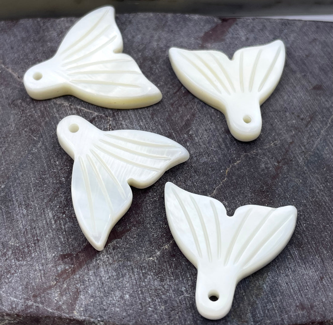 Four white carved pendant charms shaped like stylized fish or fin-like forms with a small hole, arranged on a dark textured surface.