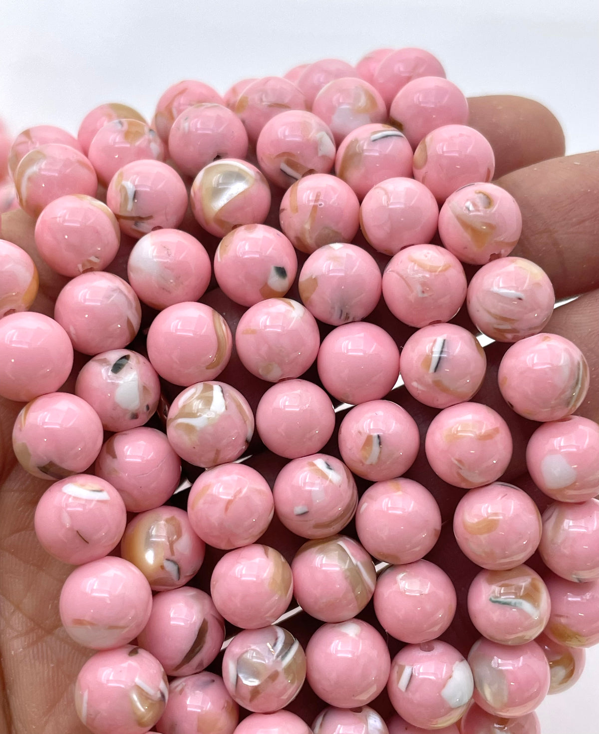 A close-up of a hand holding a dense bundle of glossy pink mop beads with white, gold, and brown swirls, set against a plain white background.