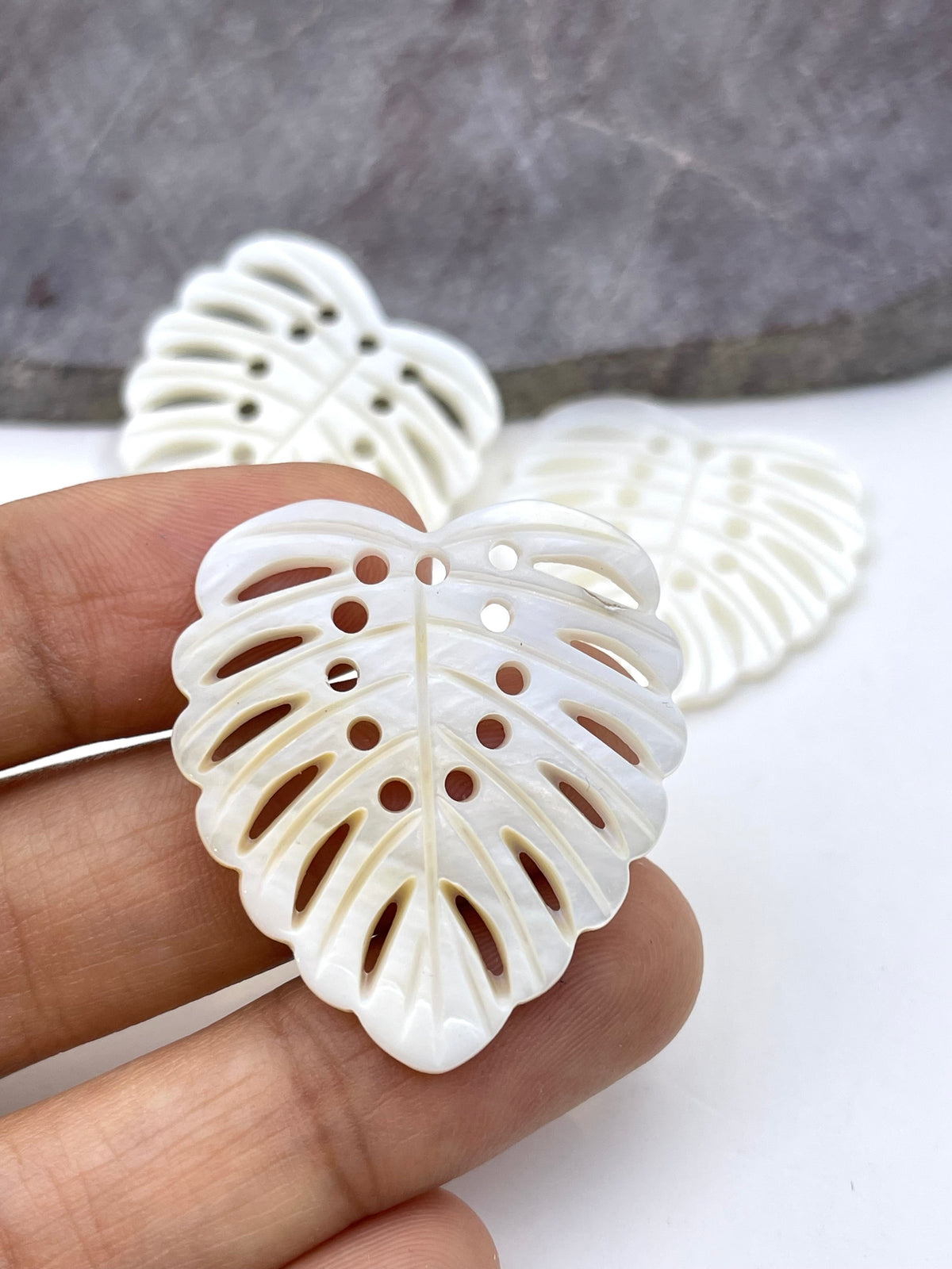A hand holding a white, leaf-shaped pendant or charm with cutout details, with two more of the same pieces blurred in the background against a white and gray surface.