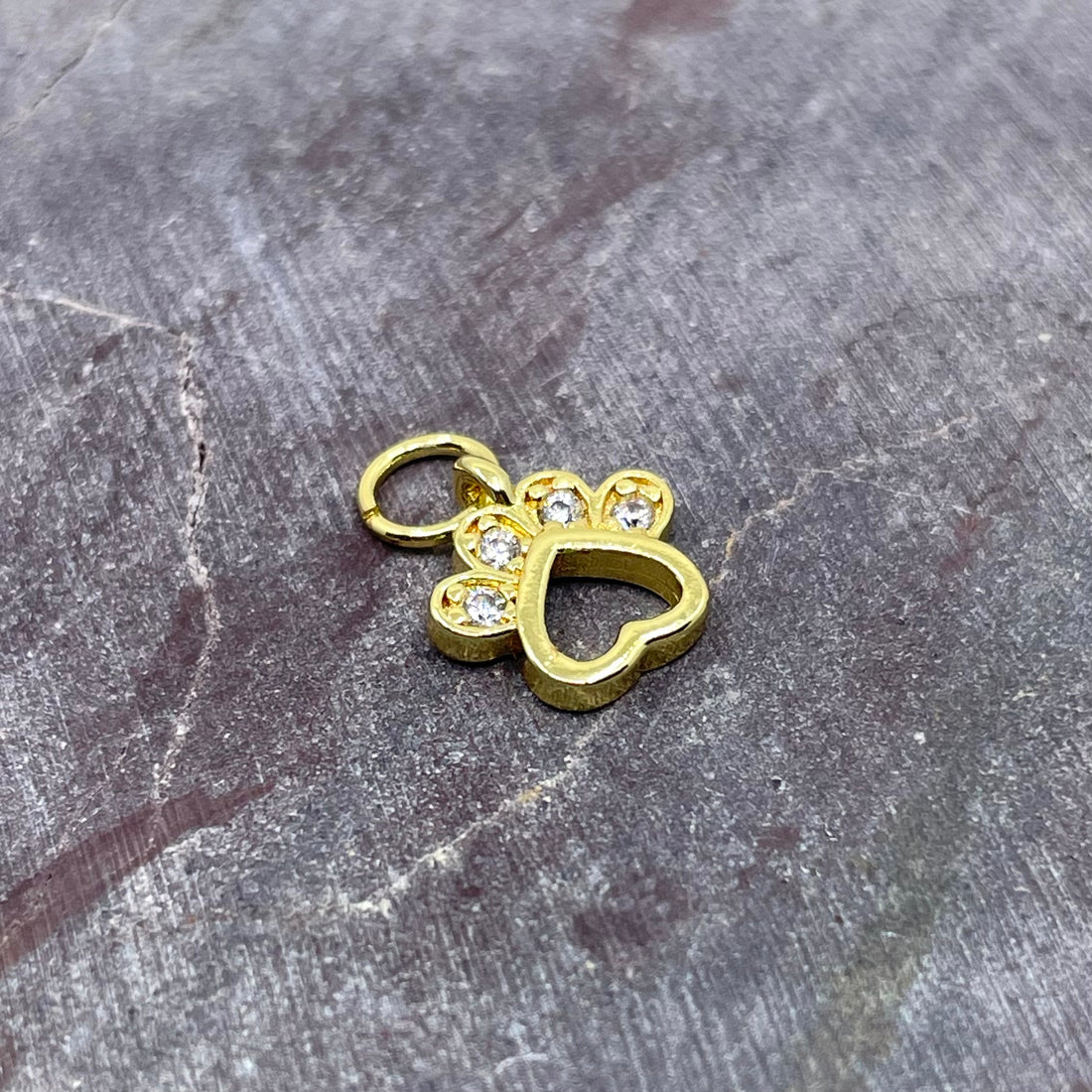 Two small paw-shaped jewelry charms with heart cutouts and clear rhinestone accents, shown on a textured gray stone surface. One charm is silver-toned and the other is gold-toned.