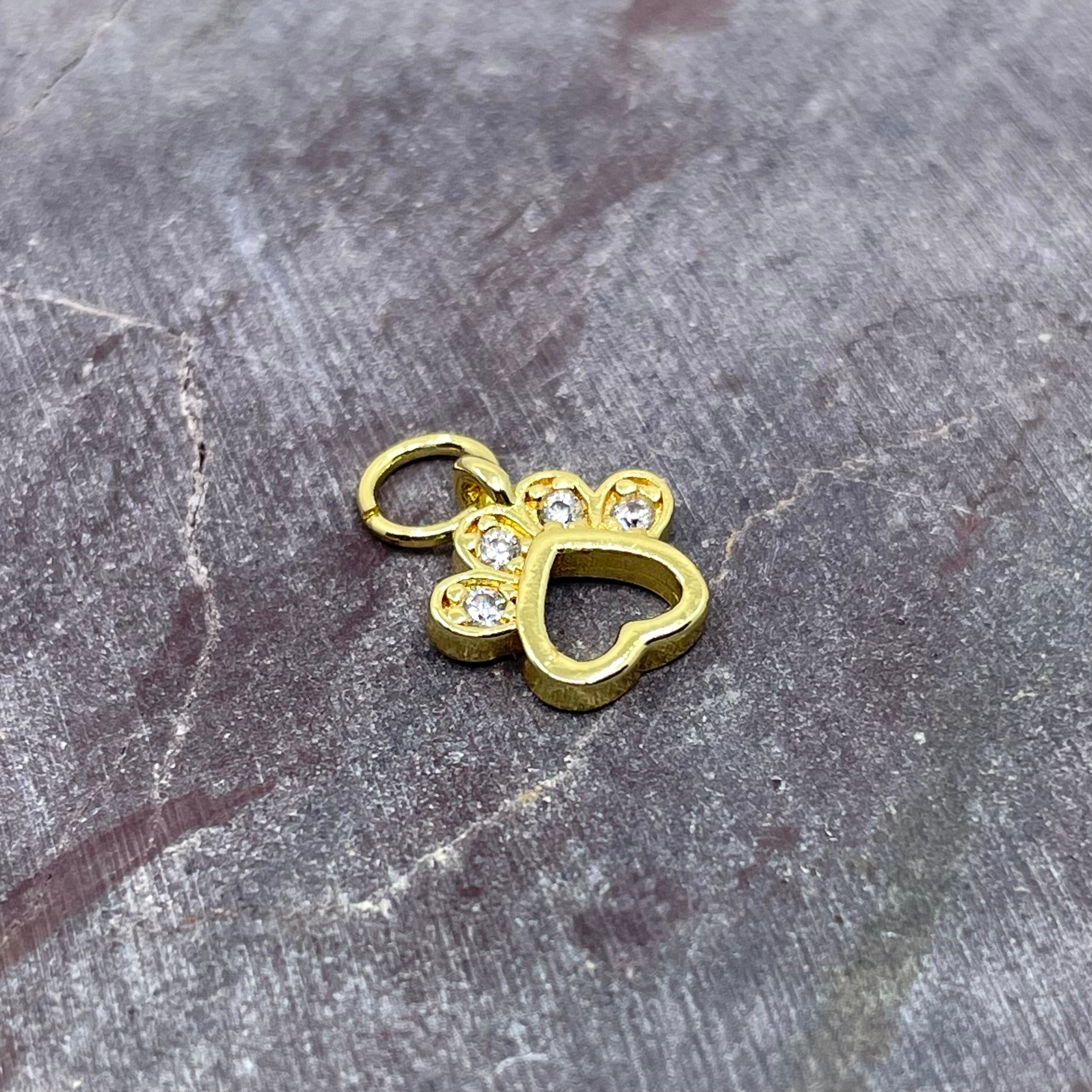 A gold-colored paw print charm with clear rhinestone accents, resting on a textured gray stone surface.