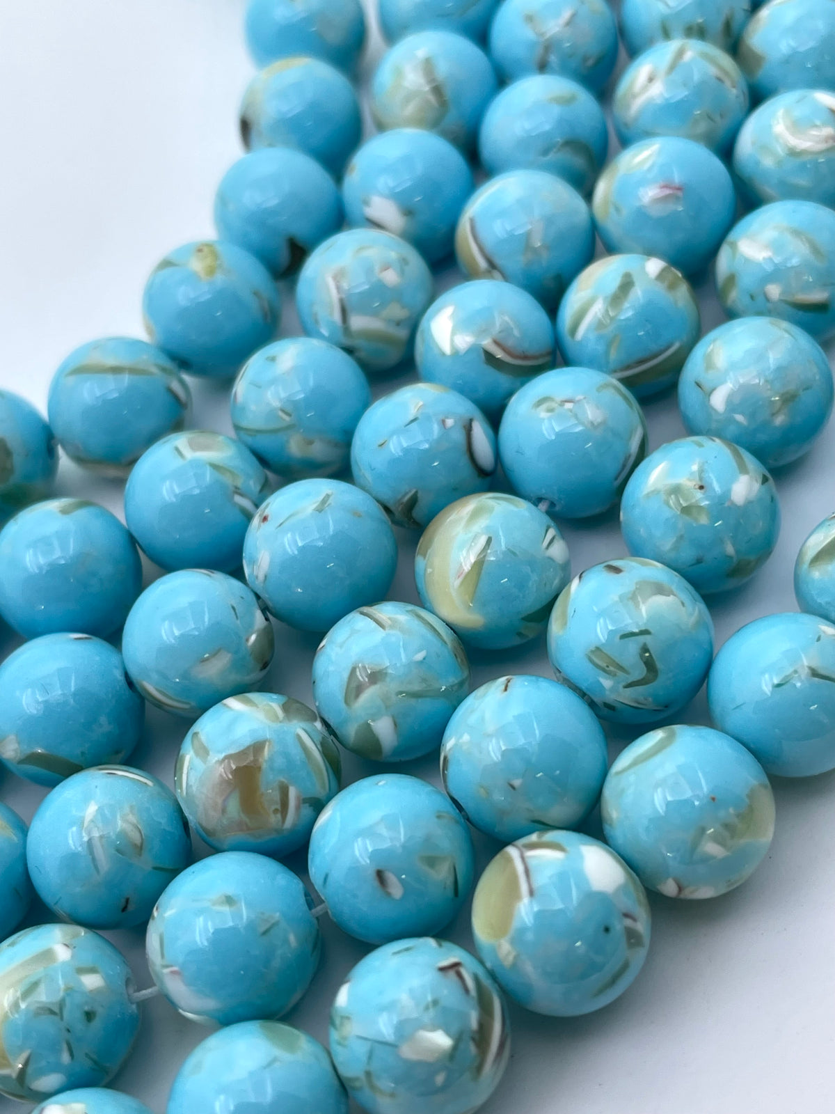 A close-up of glossy round  mop beads in a bright turquoise-blue color with marbled white and beige veining, arranged on a plain light background.