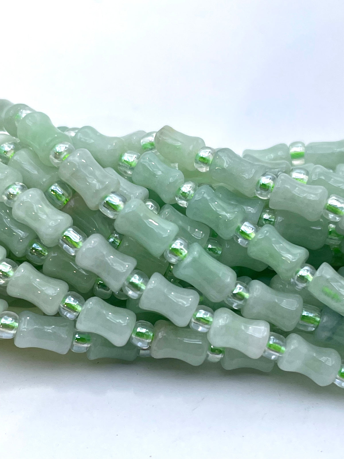 A close-up of multiple strands of light green burma jade beads, featuring cylindrical green beads separated by small clear faceted spacer beads, set against a plain white background.