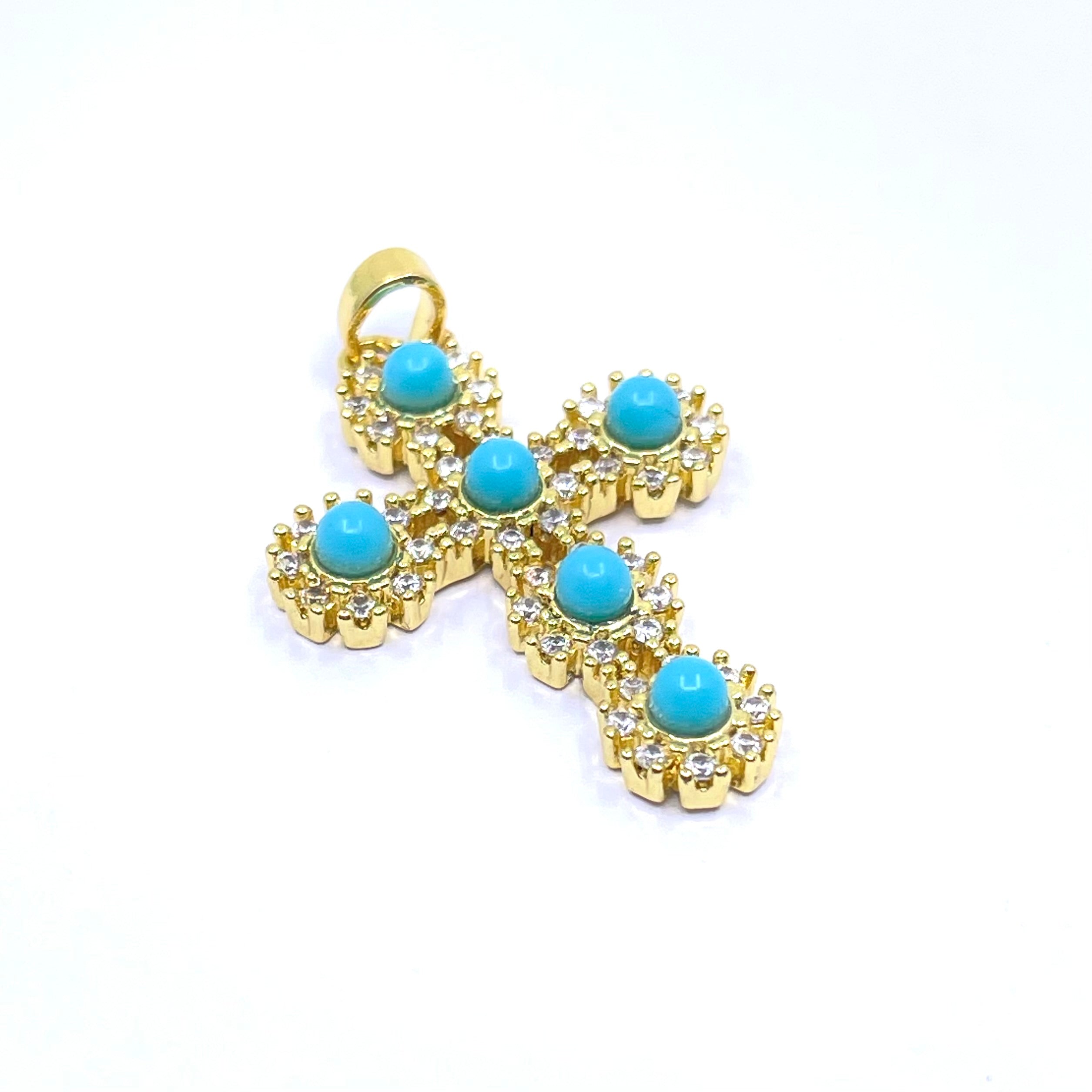 A gold cross pendant featuring SIX round turquoise-blue cabochon stones, each surrounded by a halo of small clear crystals, set against a plain white background.