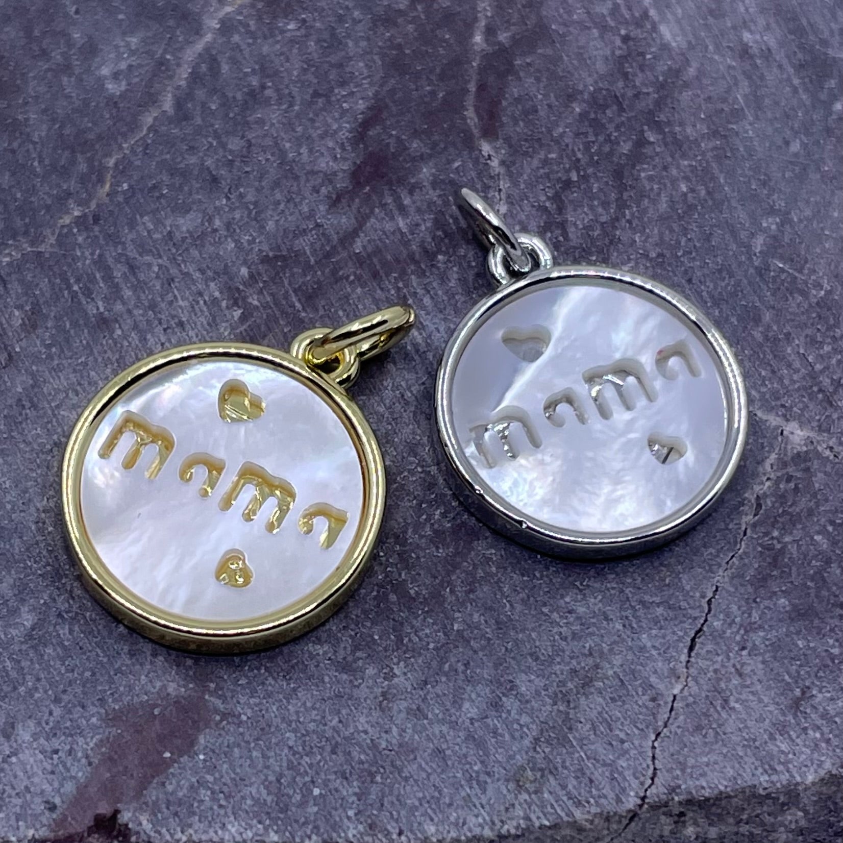 Two round pendant charms with white mother-of-pearl faces and gold lettering reading 'mama' with small heart accents, shown on a dark stone surface. One charm has a gold-toned border and bail, and the other has a silver-toned border and bail.