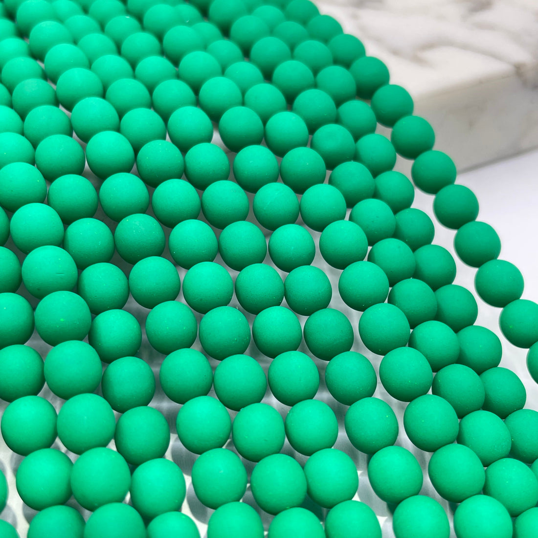 Green matte round beads arranged in parallel rows on a white surface with a blurred marble block in the background.