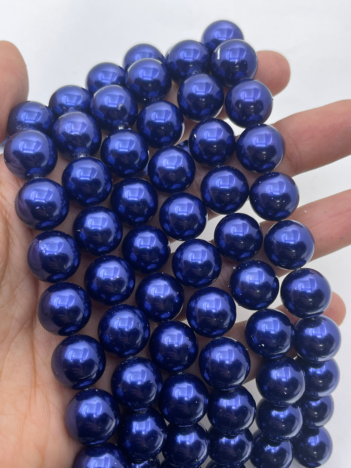 A hand holding a strand of glossy, deep blue shell pearl beads against a plain white background.
