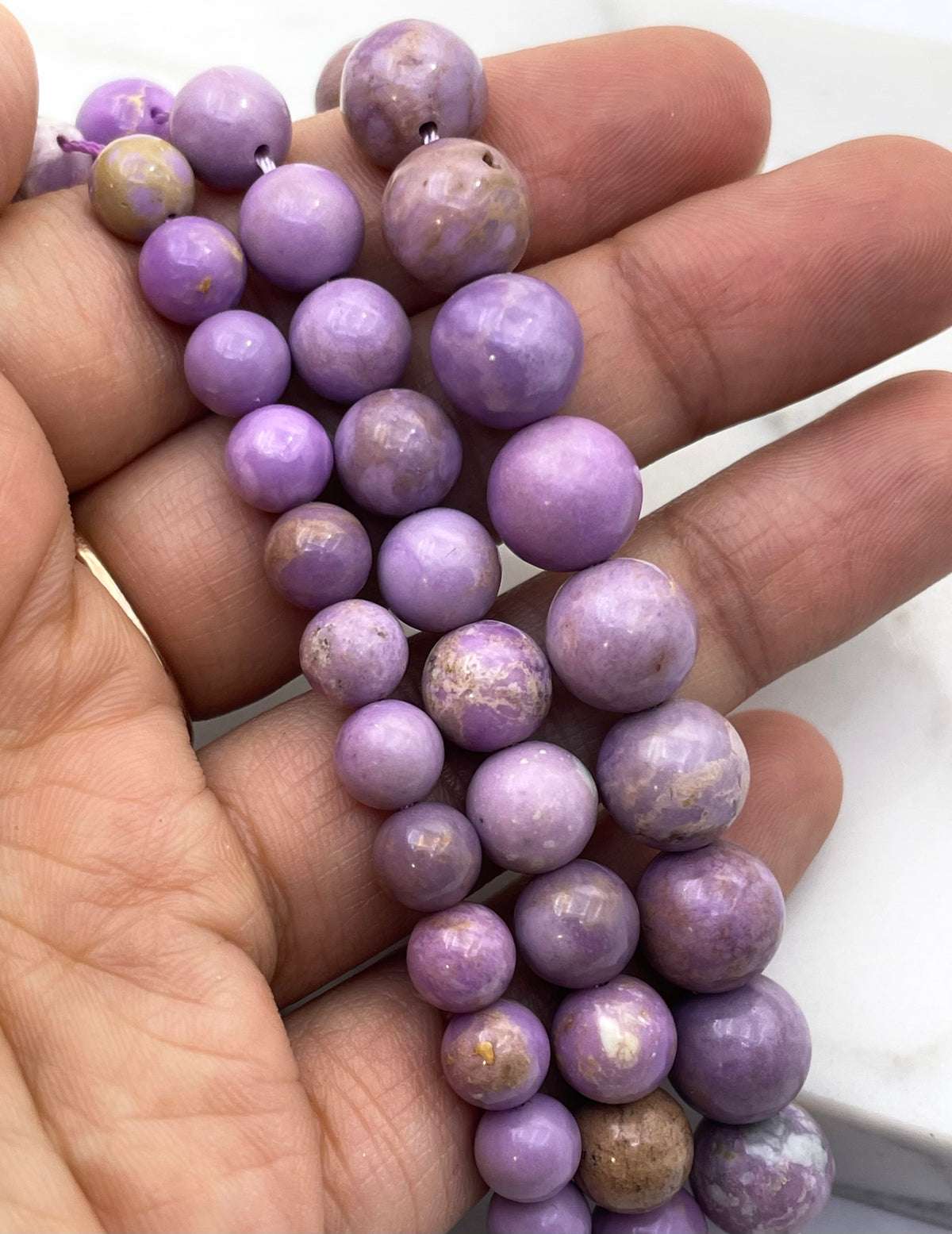 A hand holding a strand of round phosphosiderite lavender-purple beads with natural marbling and some lighter cream areas.