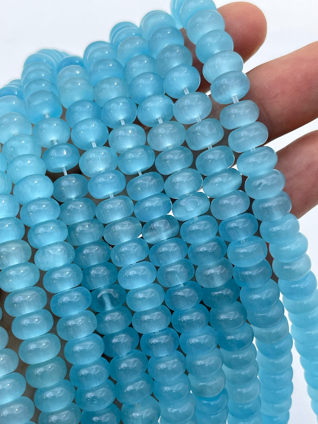 Light blue cats eye translucent donut gemstone beads strung on cords, held in a hand against a white background.