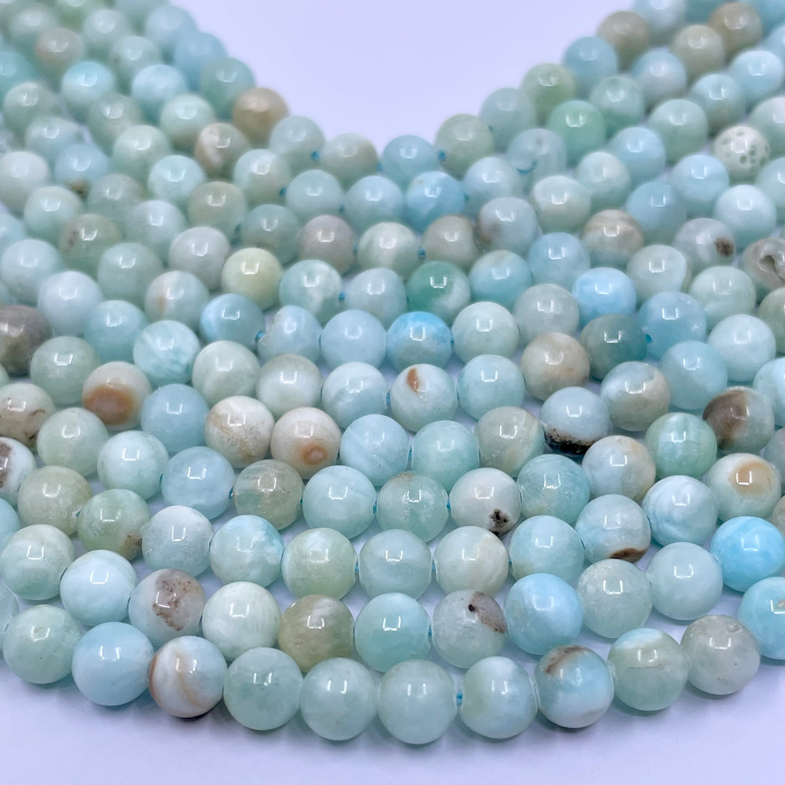 A close-up of round light blue-green hemimorphite gemstone beads with natural marbling and one striped accent bead, arranged on a white marble surface.