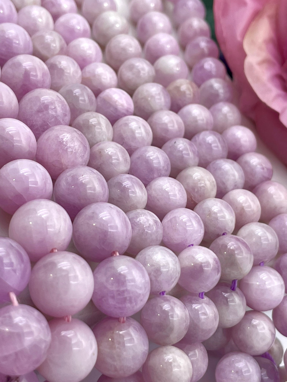 Close-up of many polished round kunzite beads in a soft lavender-pink color, arranged tightly together with a pink flower partially visible at the upper right.