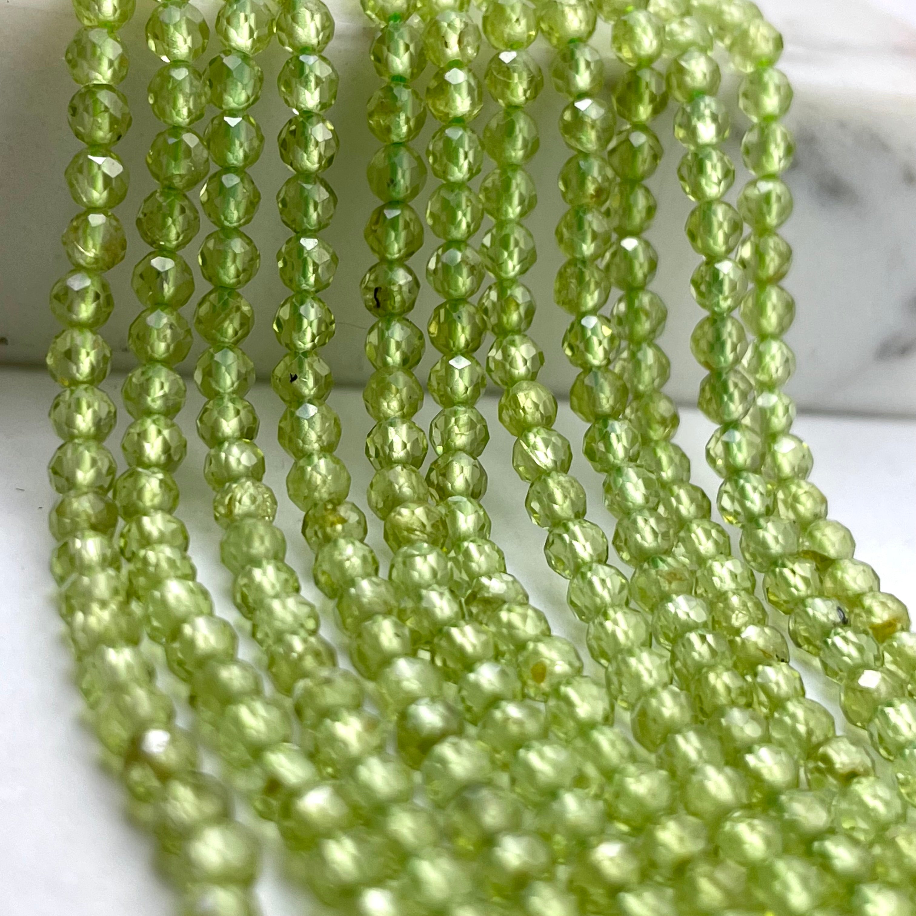 Faceted light green peridot gemstone beads arranged in multiple strands, photographed close-up against a light marble-like background.