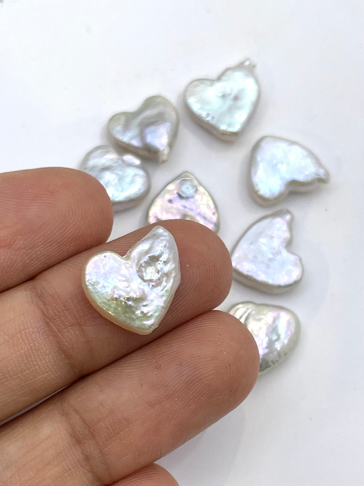 A hand holding a small, irregularly shaped iridescent white heart-shaped bead or charm, with several similar heart-shaped pieces scattered on a plain white surface in the background.