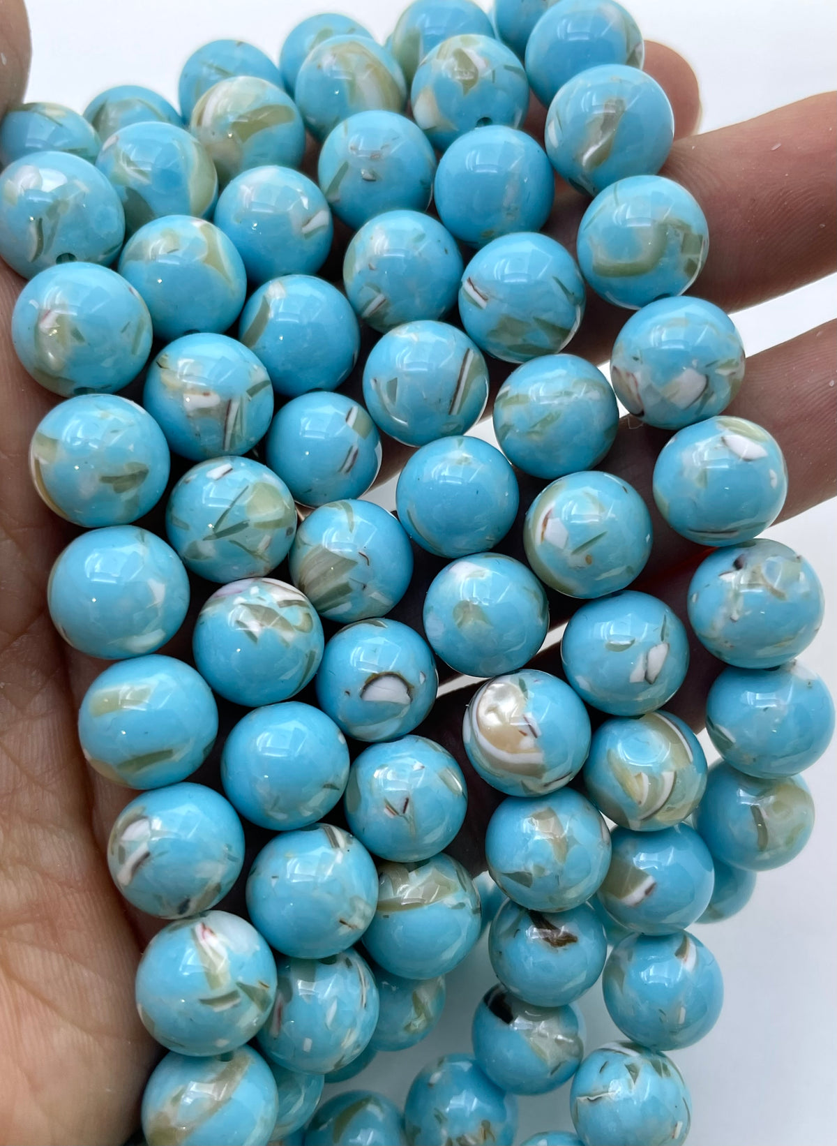A close-up of a hand holding strands of round turquoise-blue mop beads with white and light brown marbled inclusions, set against a plain white background.