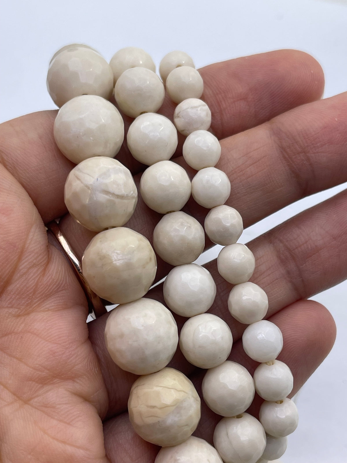 A close-up of a hand holding three strands of white, faceted fossil stone round beads in various sizes against a plain white background.