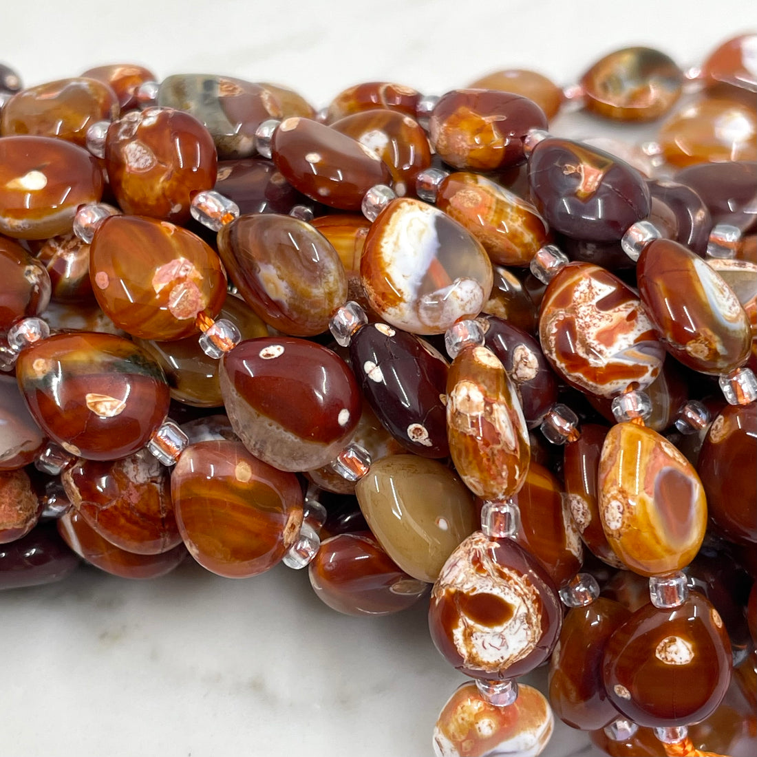 A close-up of a strand of polished TERRACOTTA beads in earthy brown, amber, and reddish tones, separated by small silver spacer beads, resting on a light marble surface.