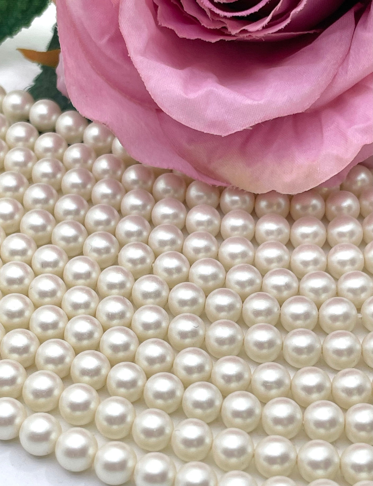 A close-up of many small round ivory shell pearls beads arranged in rows, with a pink rose placed on top in the upper right corner.