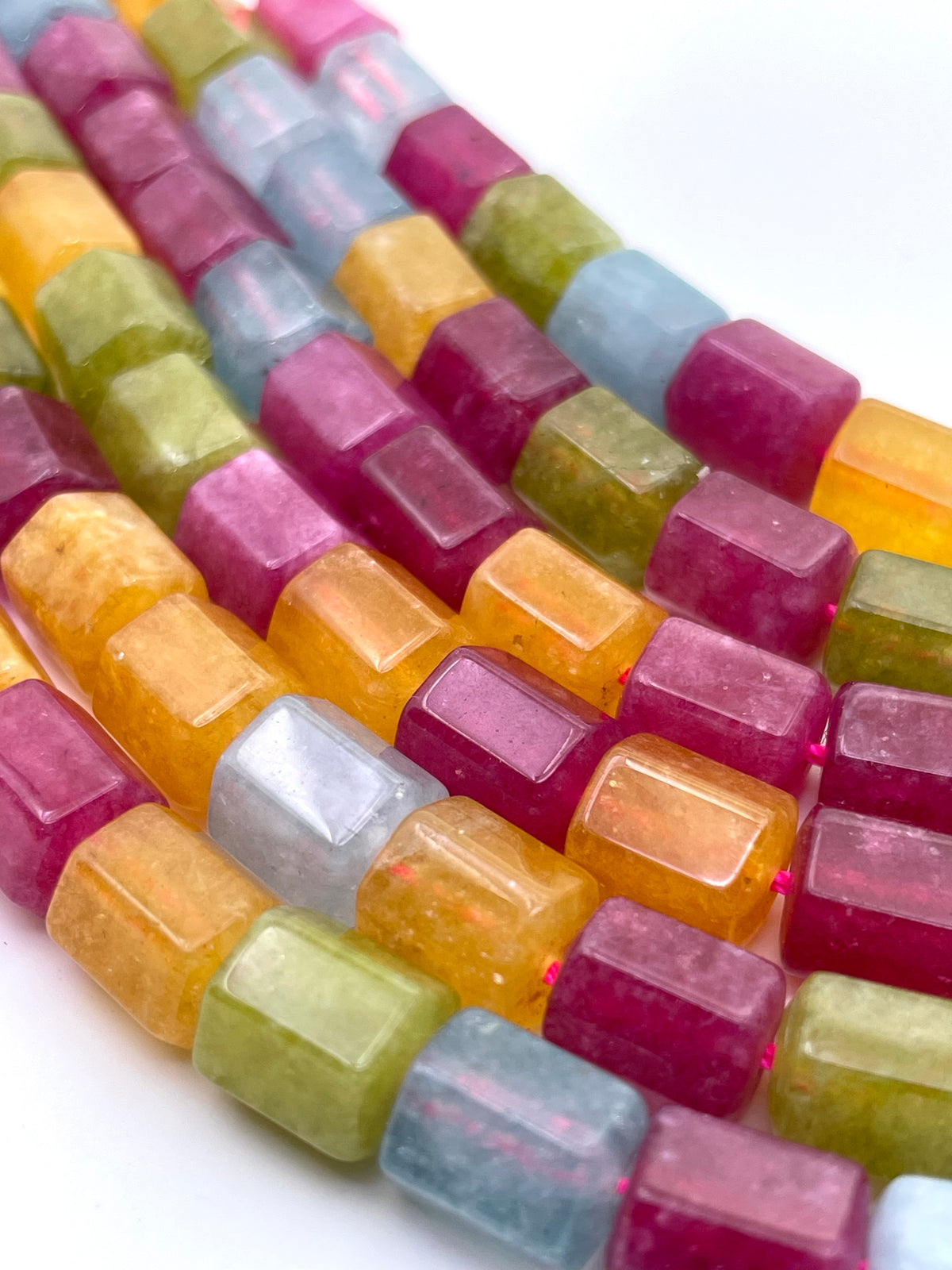 Close-up of multiple strands of faceted rectangular jade gemstone beads in assorted colors including pink, yellow, green, blue, and clear, arranged diagonally on a white background.