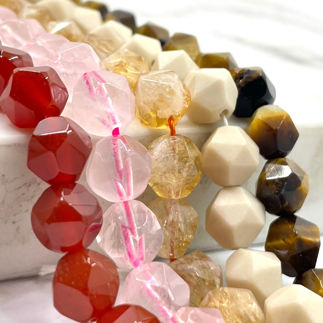 Close-up of faceted gemstone beads draped together on a white surface, including carnelian, rose quartz, citrine, fossil stone and tiger eye