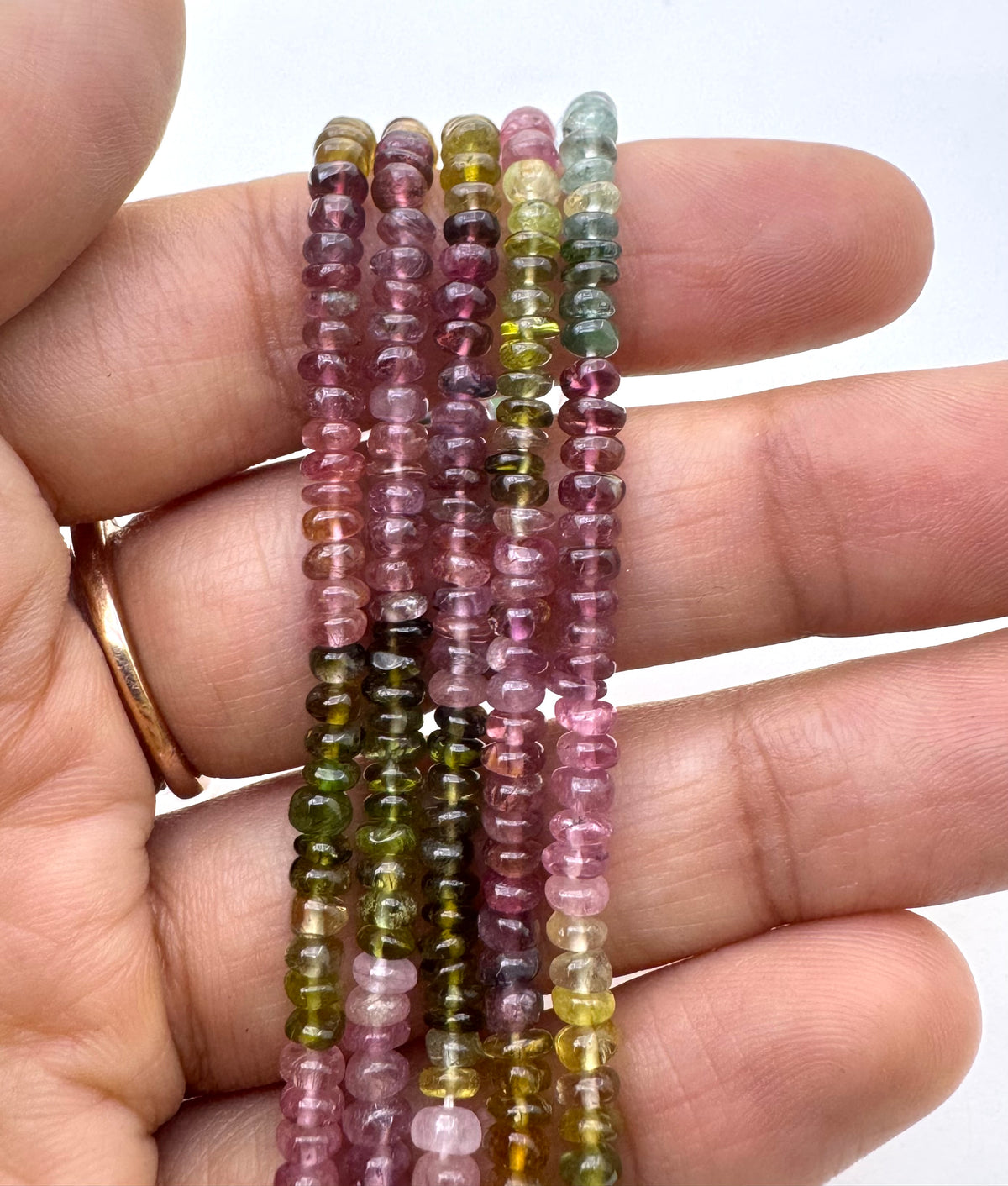 A close-up of a hand holding several strands of small disc shape mix tourmaline beads in pink, green, purple, and yellow tones against a plain light background.