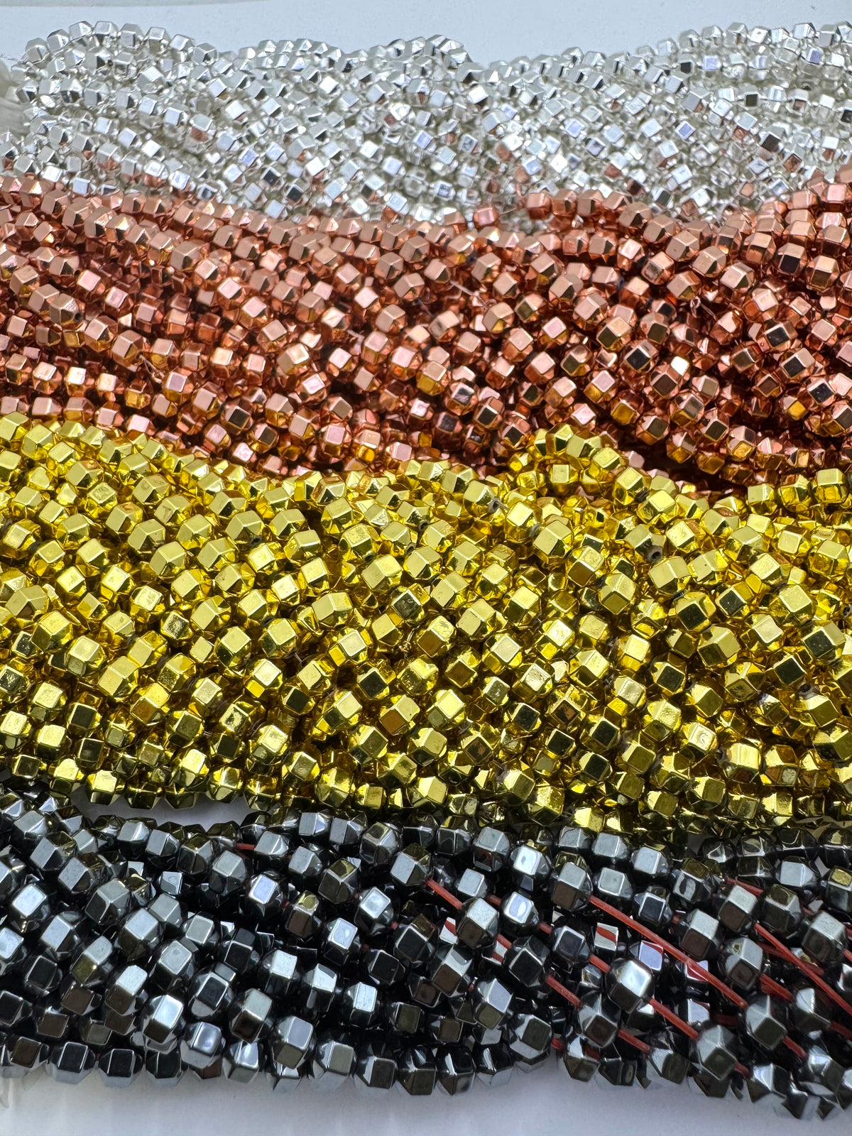 Multiple strands of metallic hematite beads arranged in horizontal rows, showing silver, rose gold, yellow gold, and gunmetal gray colors.