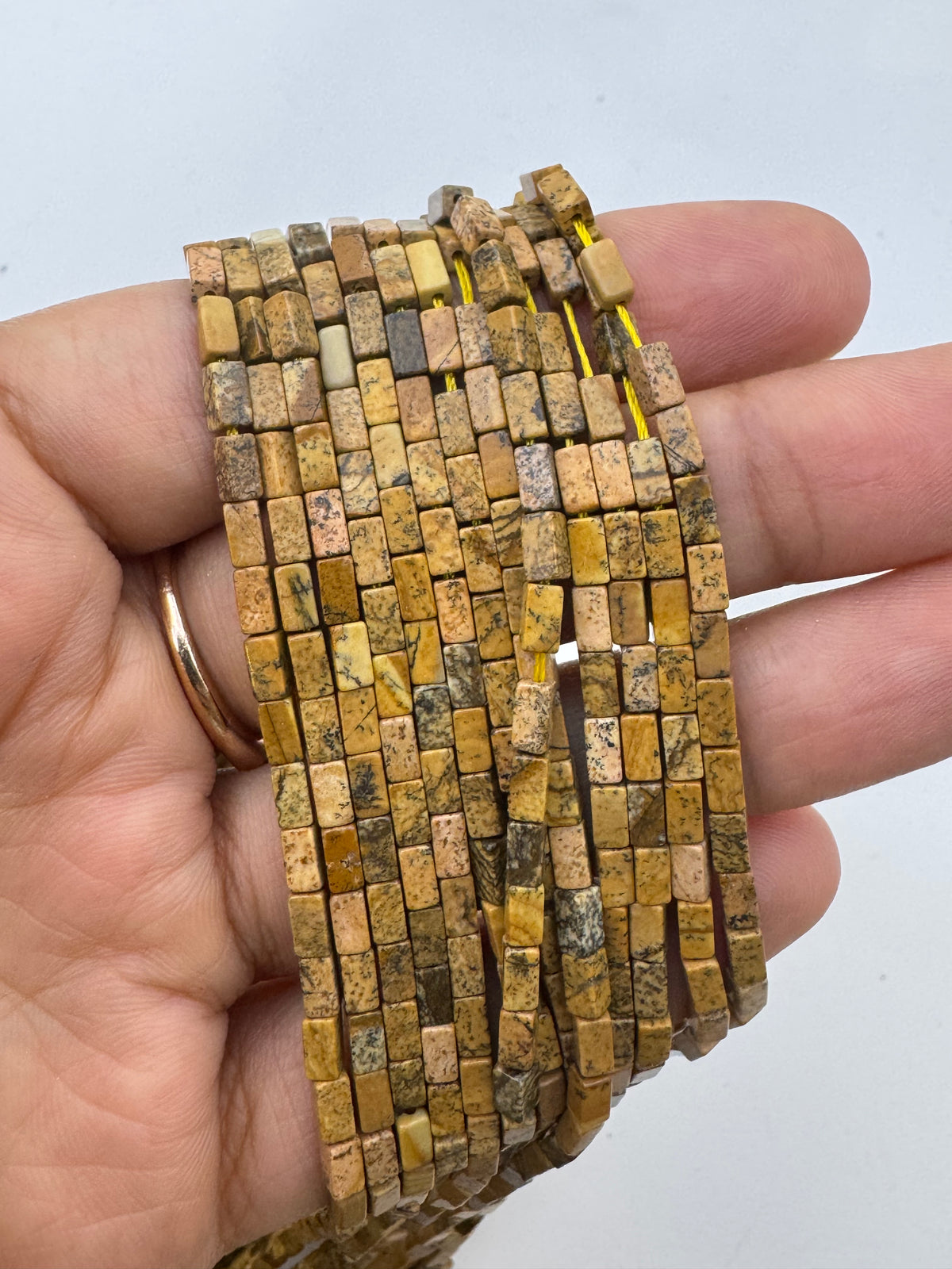 A hand holding a bundle of multiple strands of small rectangular picture jasper stone beads in earthy yellow, brown, and beige colors, with a gold ring on one finger, set against a plain light background.