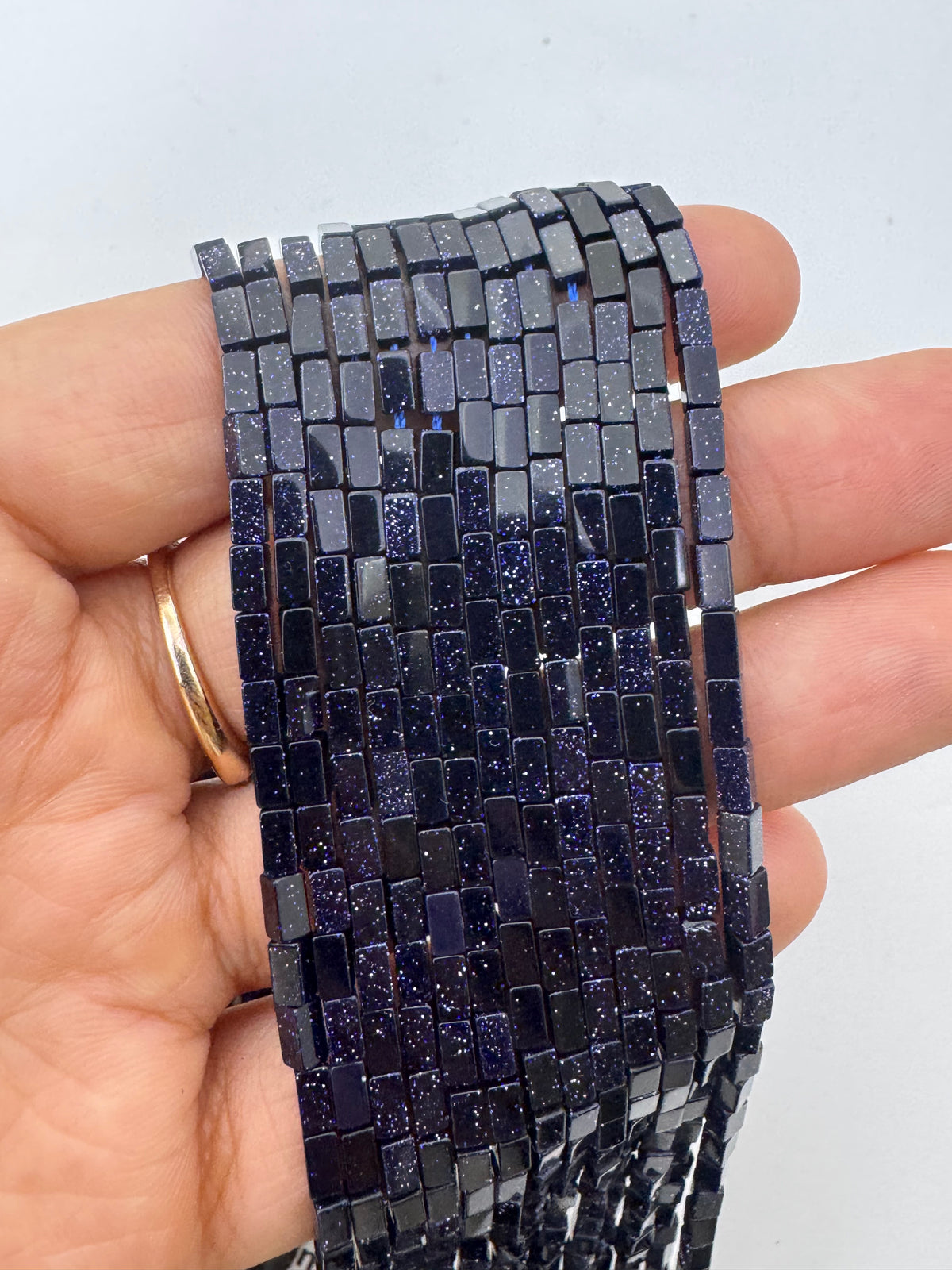 A hand holding a long strand of dark rectangular blue sandstone beads with a glittery, metallic finish, set against a plain light background.