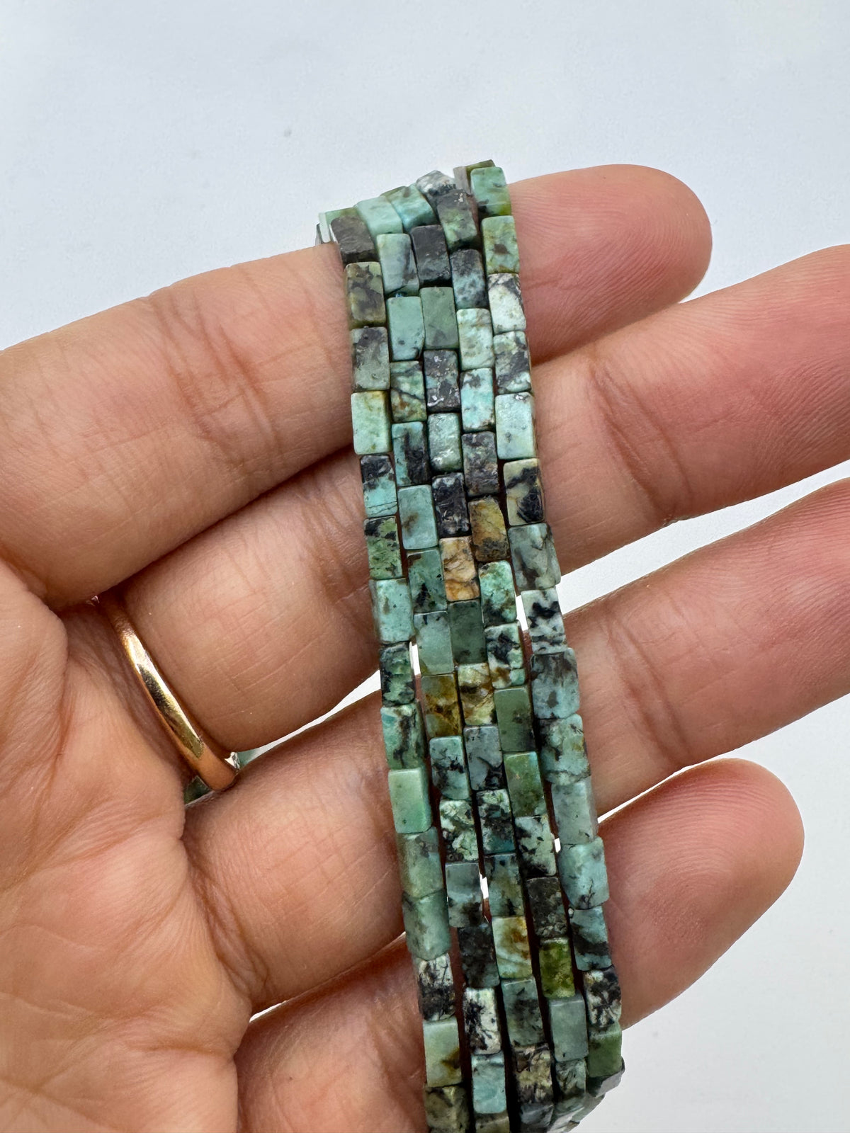 A close-up of a hand holding a multi-strand bracelet made of small rectangular african turquoise green stone beads with black and brown matrix patterns, against a plain light background.