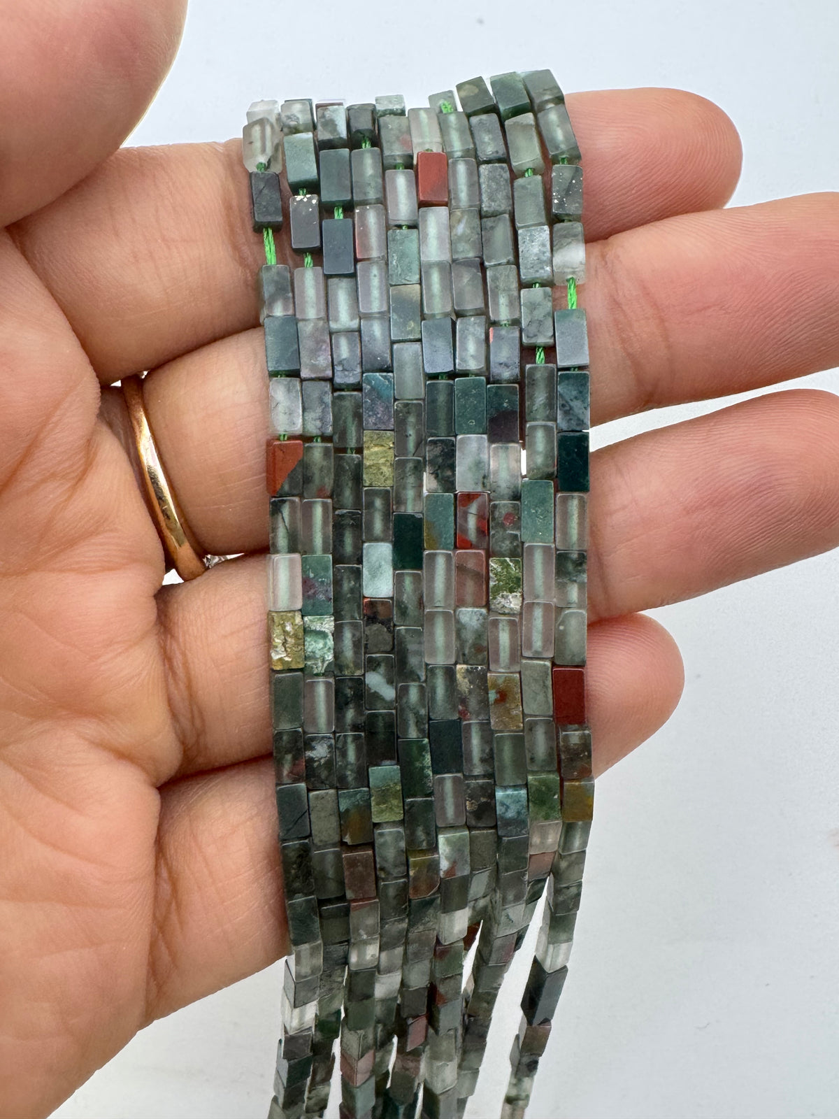A hand holding a bundle of multi-strand rectangular bloodstone bead strands made from polished stone chips in earthy green, gray, brown, and reddish tones, against a plain light background.