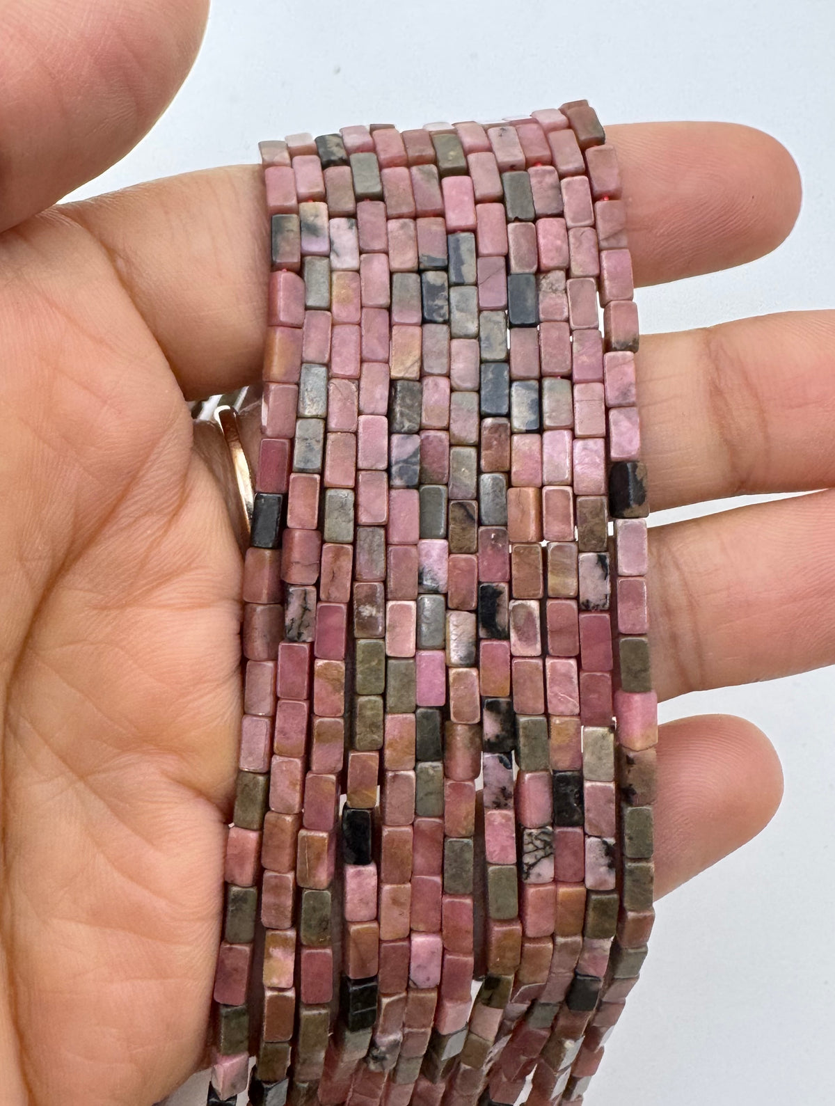 A hand holding a long strand of small rectangular rhodonite beads in pink, mauve, brown, gray, and black tones against a plain light background.