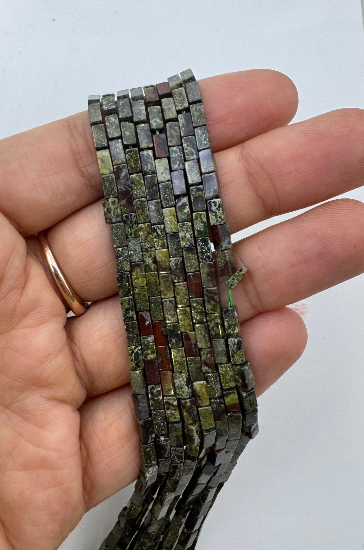 A hand holding a strand of rectangular dragon stone gemstone beads, showing their varied earthy colors and textures against a plain white background.