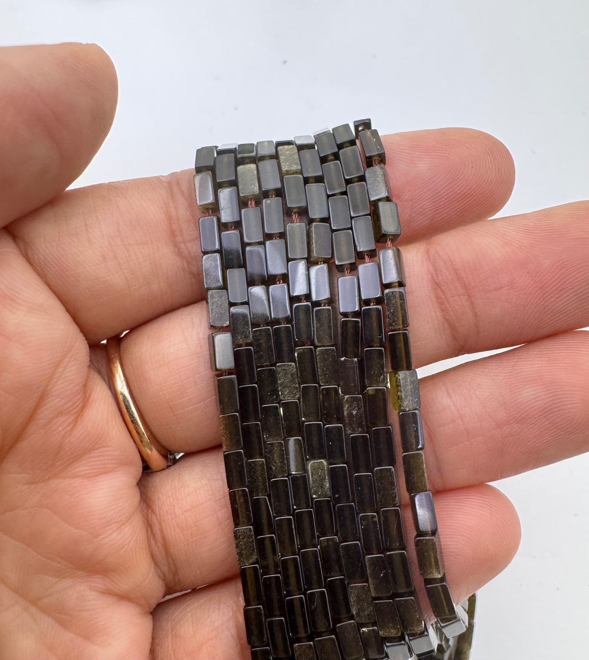 A hand holding a strand of small rectangular gold obsidian dark gray to black beads, with a gold ring visible on one finger, against a plain light background.