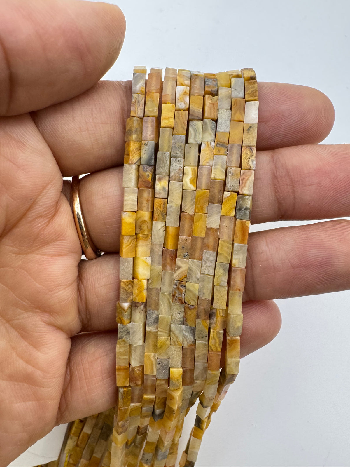 A hand holding a strand of small rectangular crazy lace agate stone beads in warm yellow, tan, cream, and brown marbled tones against a plain light background.
