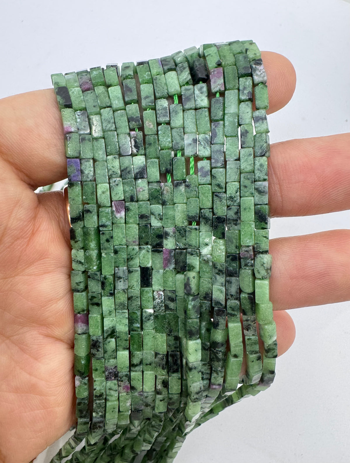 A hand holding a long strand of small rectangular ruby zoisite stone beads with natural marbling and some purple inclusions, against a plain light background.