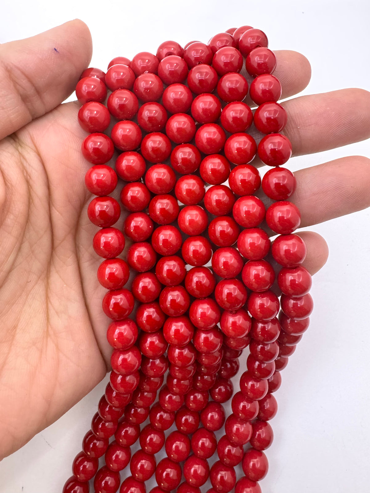 A hand holding a strand of glossy, round red shell pearl beads against a plain white background.